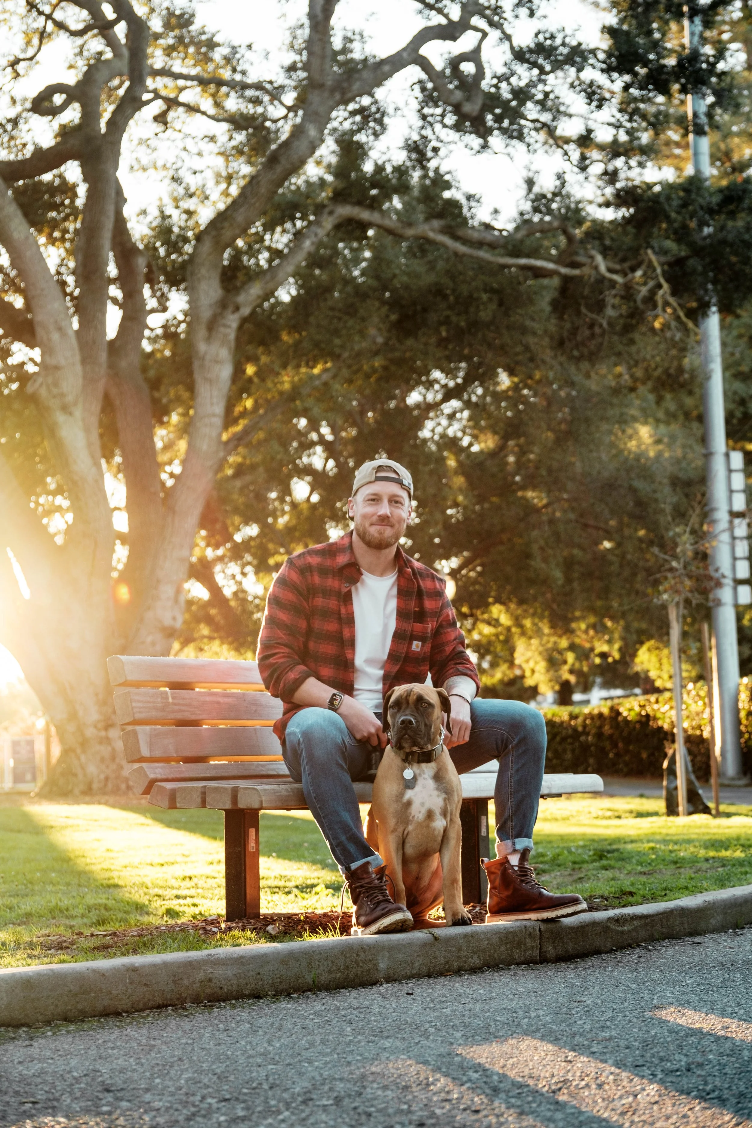 A man sitting on a park bench with his dog during sunset, trees in the background, wearing casual clothing.