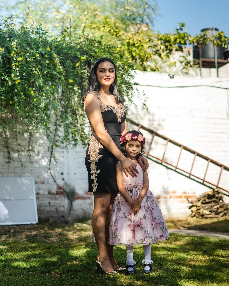 A woman and a young girl standing outdoors on grass, with green foliage and a white brick wall behind them. The woman wears a black dress with gold lace accents, and the girl wears a pink floral dress with a flower crown. They are smiling, with the w