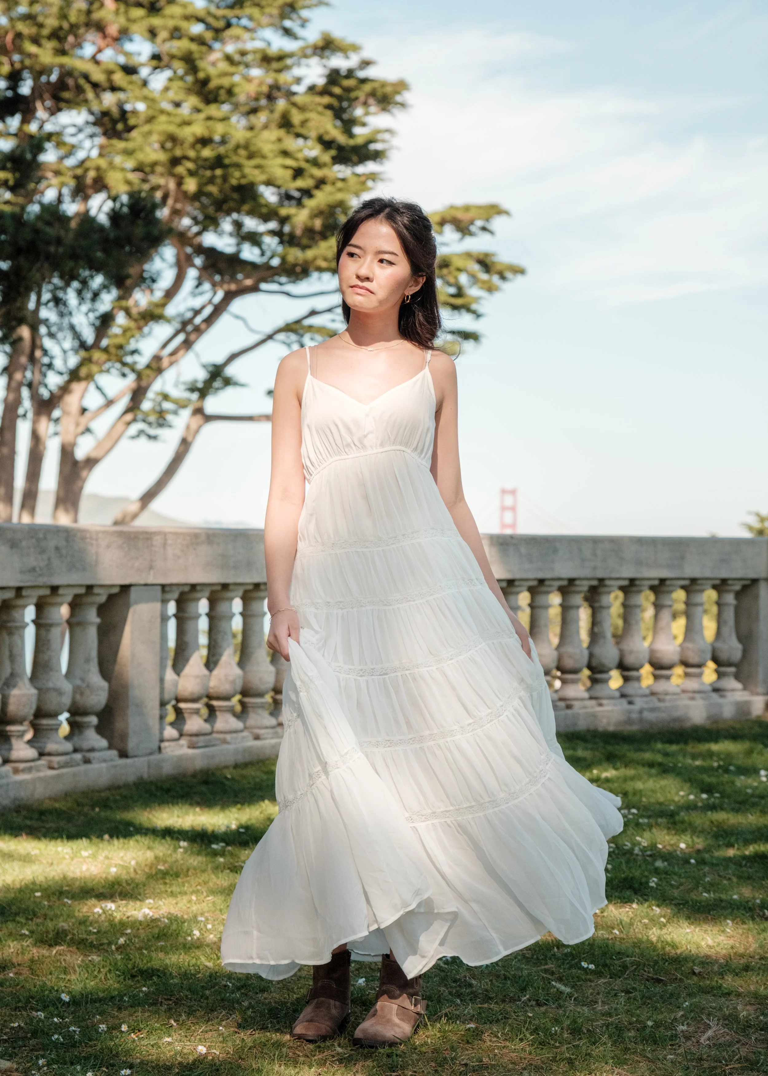 A young woman in a white flowing dress standing outdoors on grass, with a railing, trees, and the Golden Gate Bridge in the background, during daytime.