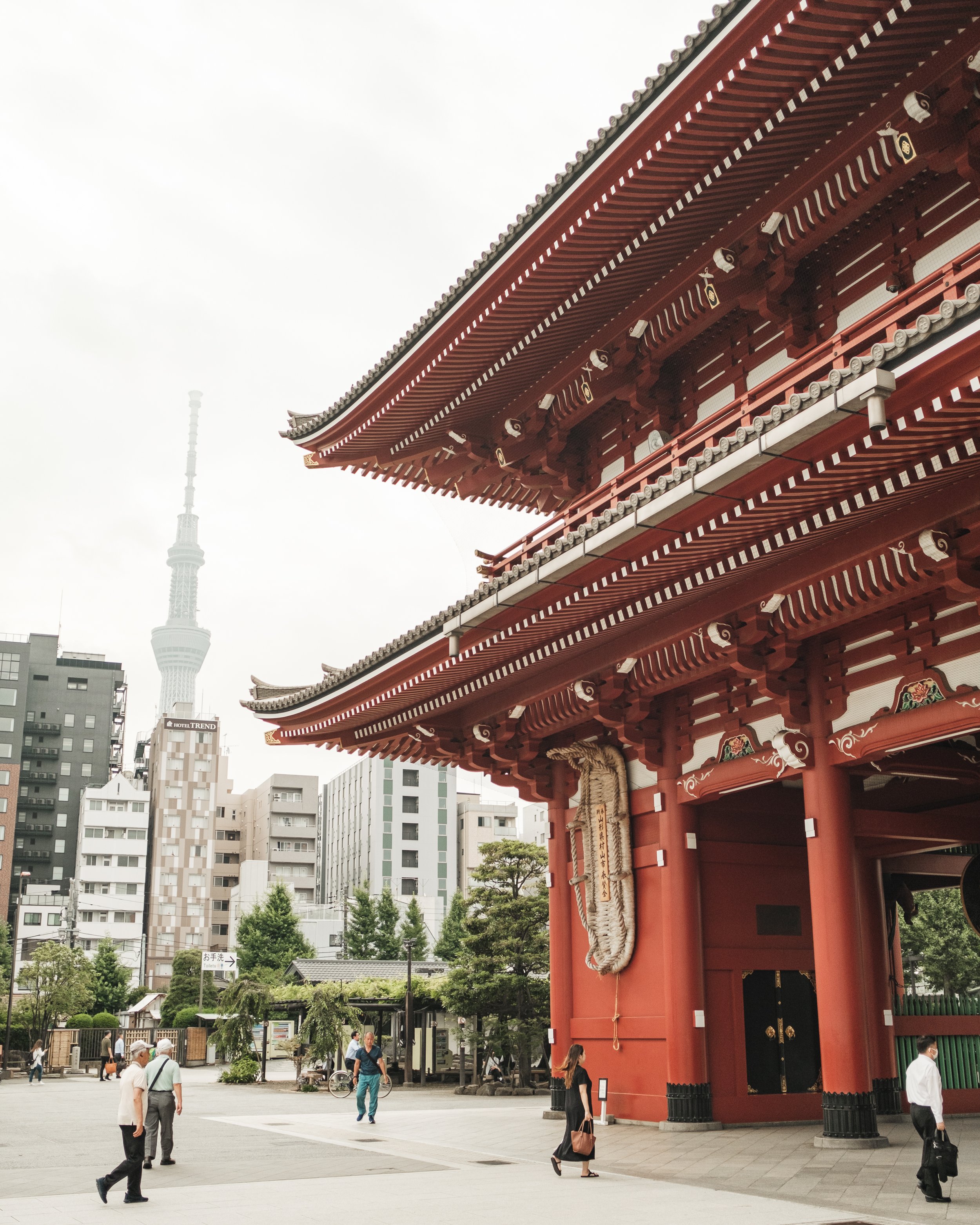 People walking past a traditional Japanese temple gate with high-rise buildings and Tokyo Skytree tower in the background.