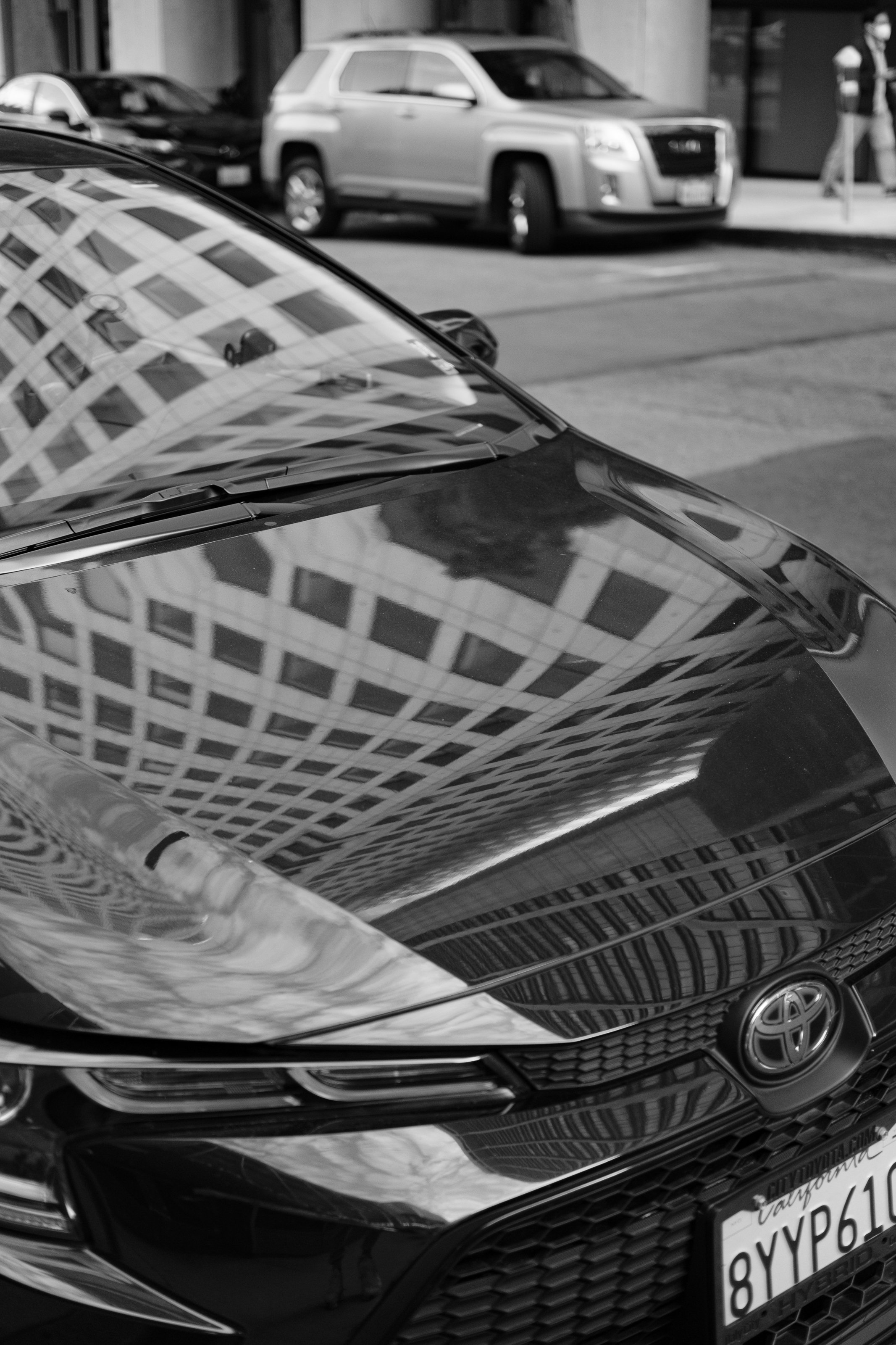 Reflected image of a modern car on a building facade with other vehicles and pedestrians on the street in the background.