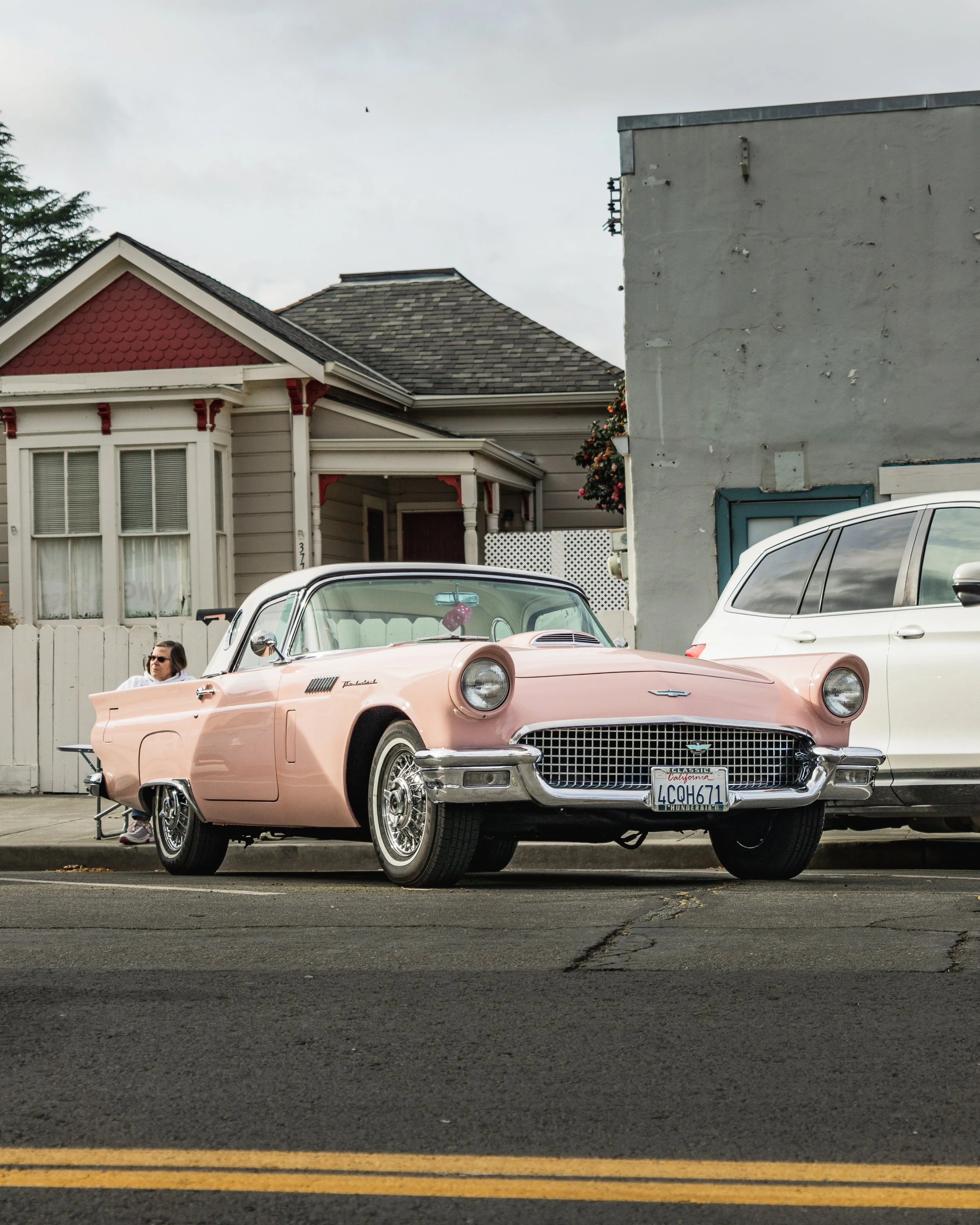 A pink classic Ford Thunderbird car is parked on the street beside a white modern SUV. There is a woman sitting on a bench near the car, with houses and a cloudy sky in the background.