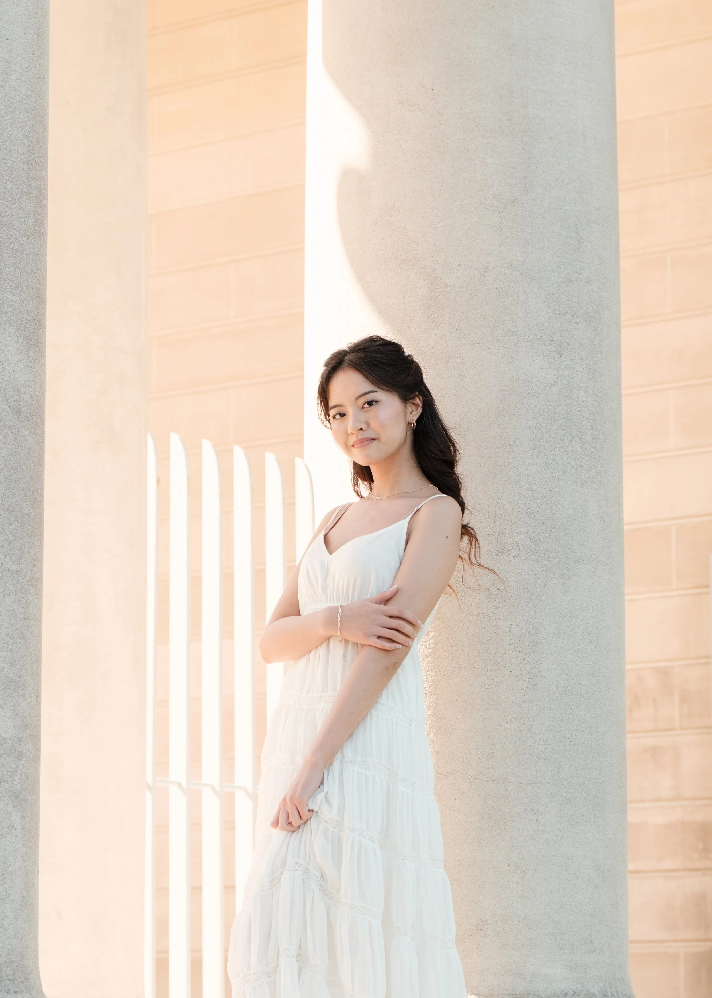 A young woman in a white sleeveless dress standing next to large columns outside, with a modern building and a white fence in the background.