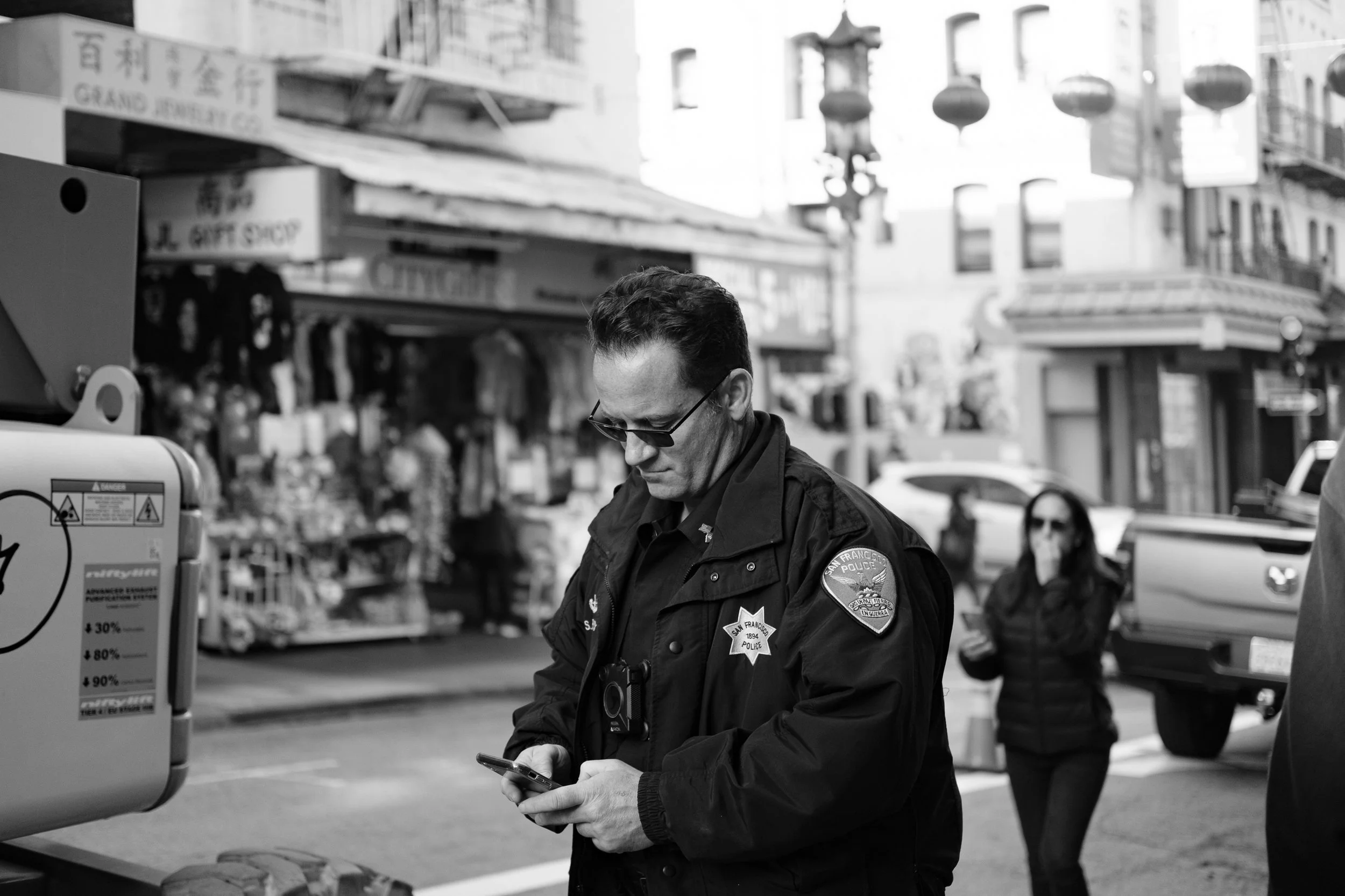 A San Francisco police officer is looking at his smartphone on a city street with shops and lanterns in the background. Another person is walking behind him in the distance.