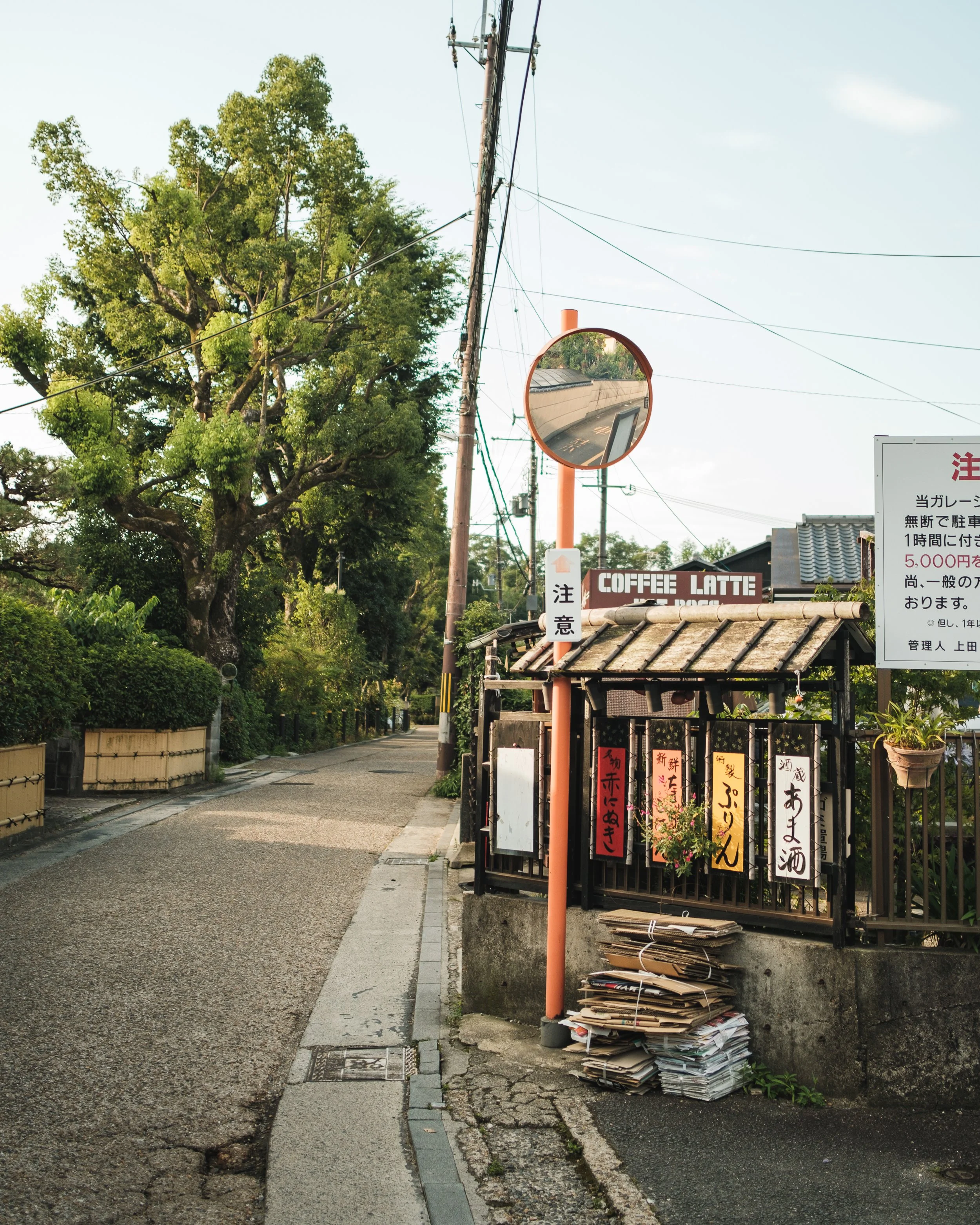 A quiet street with a small roadside coffee shop, electrical poles, a convex mirror, and a pile of newspapers and cardboard on the sidewalk. Trees line the street, and clear skies are above.