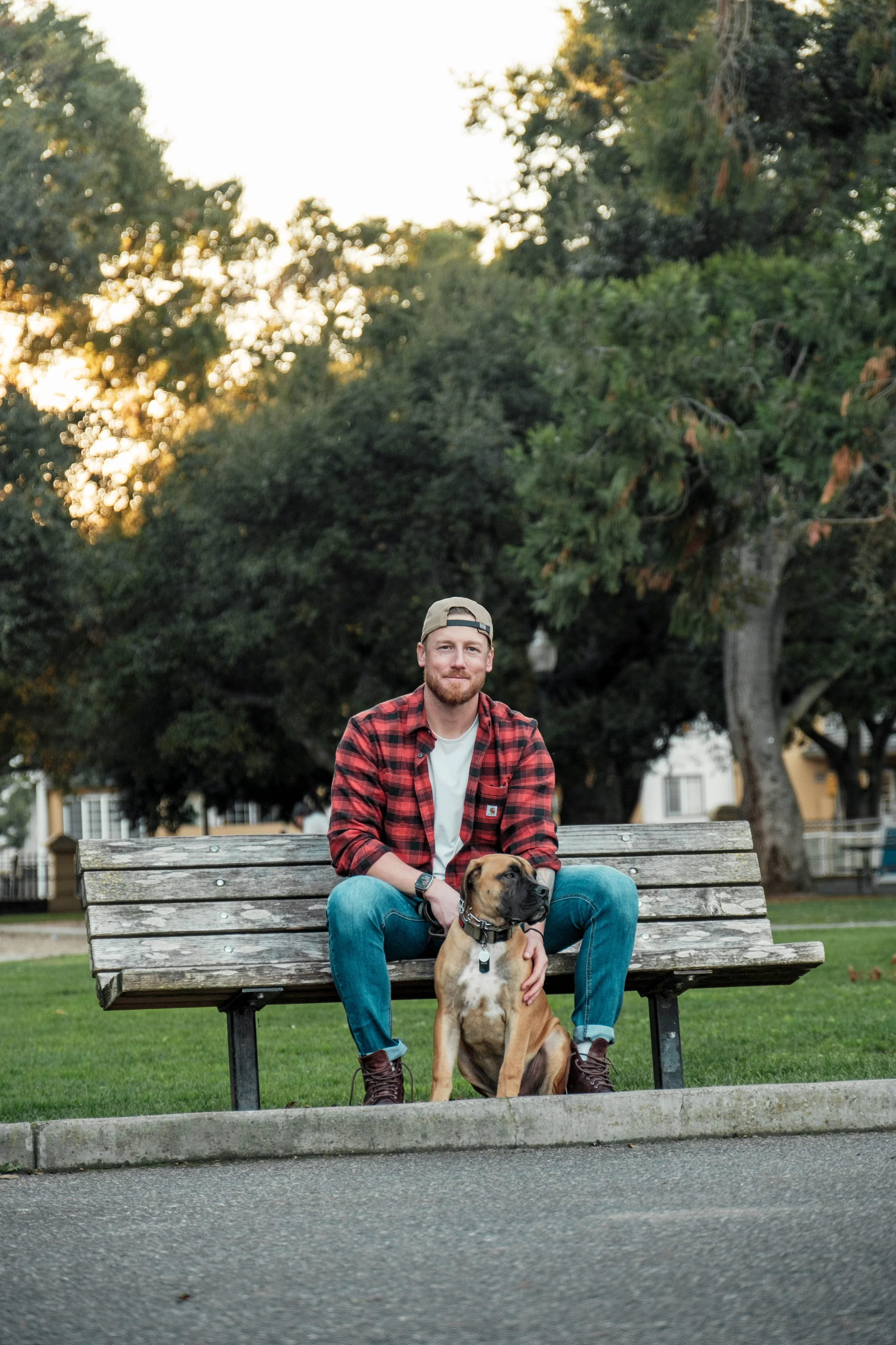 A man in a red plaid shirt and jeans sitting on a park bench with a brown dog, surrounded by green trees during sunset.