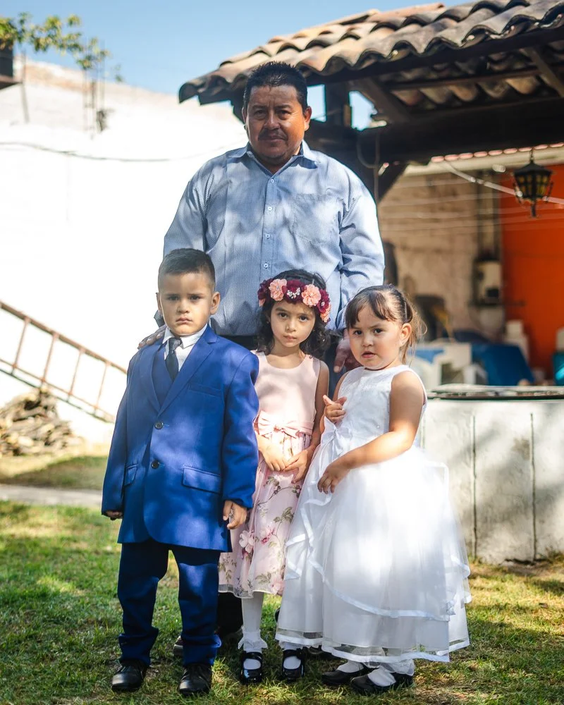 A man with three children standing outdoors in a backyard, with a house and a tree in the background. The man is wearing a light blue shirt, and the children are dressed in formal clothes: a boy in a blue suit, a girl in a floral dress with a flower 