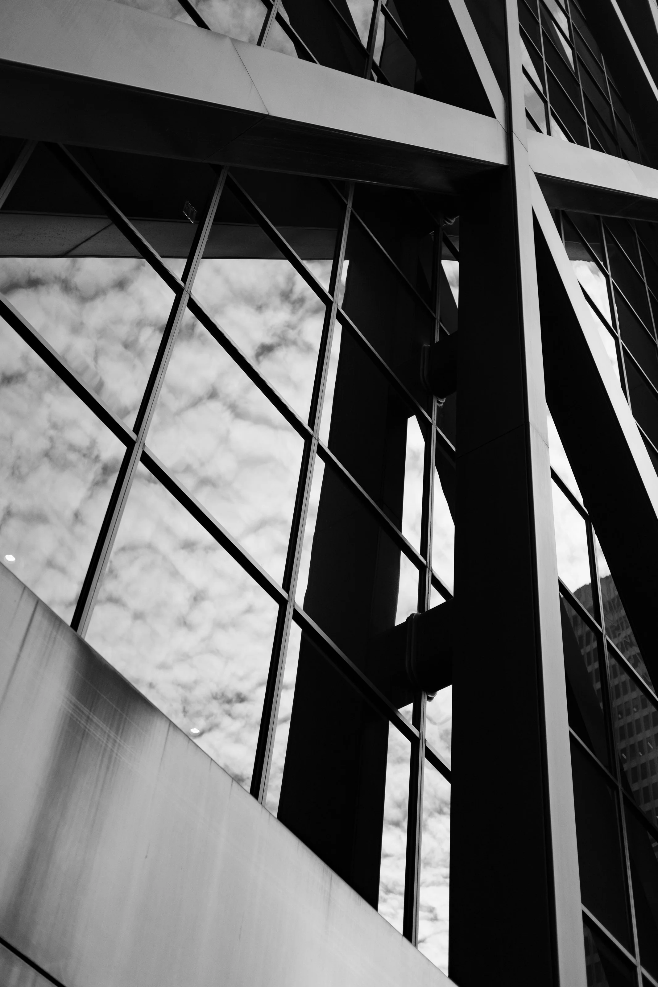 Black and white photo of a modern building with glass windows and structural framework reflecting the sky with scattered clouds.