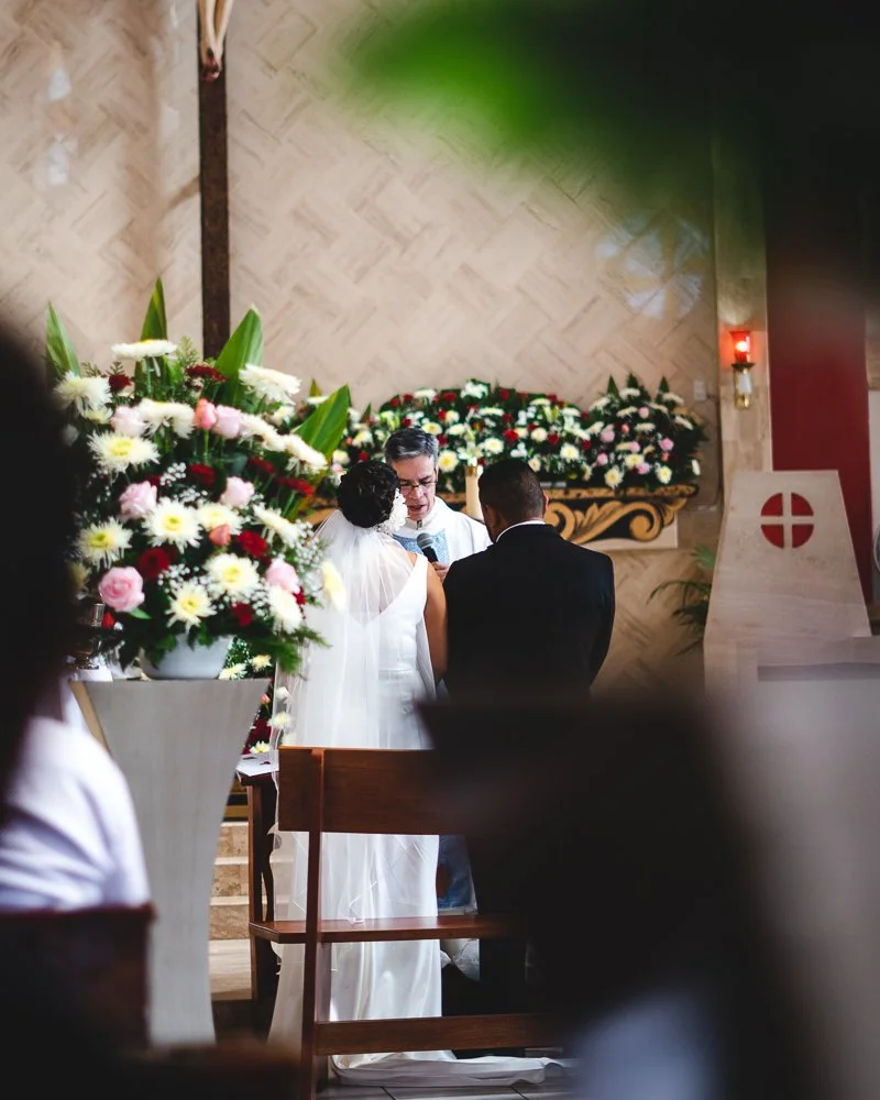 A wedding ceremony taking place inside a church with a couple standing before a priest, surrounded by large flower arrangements.