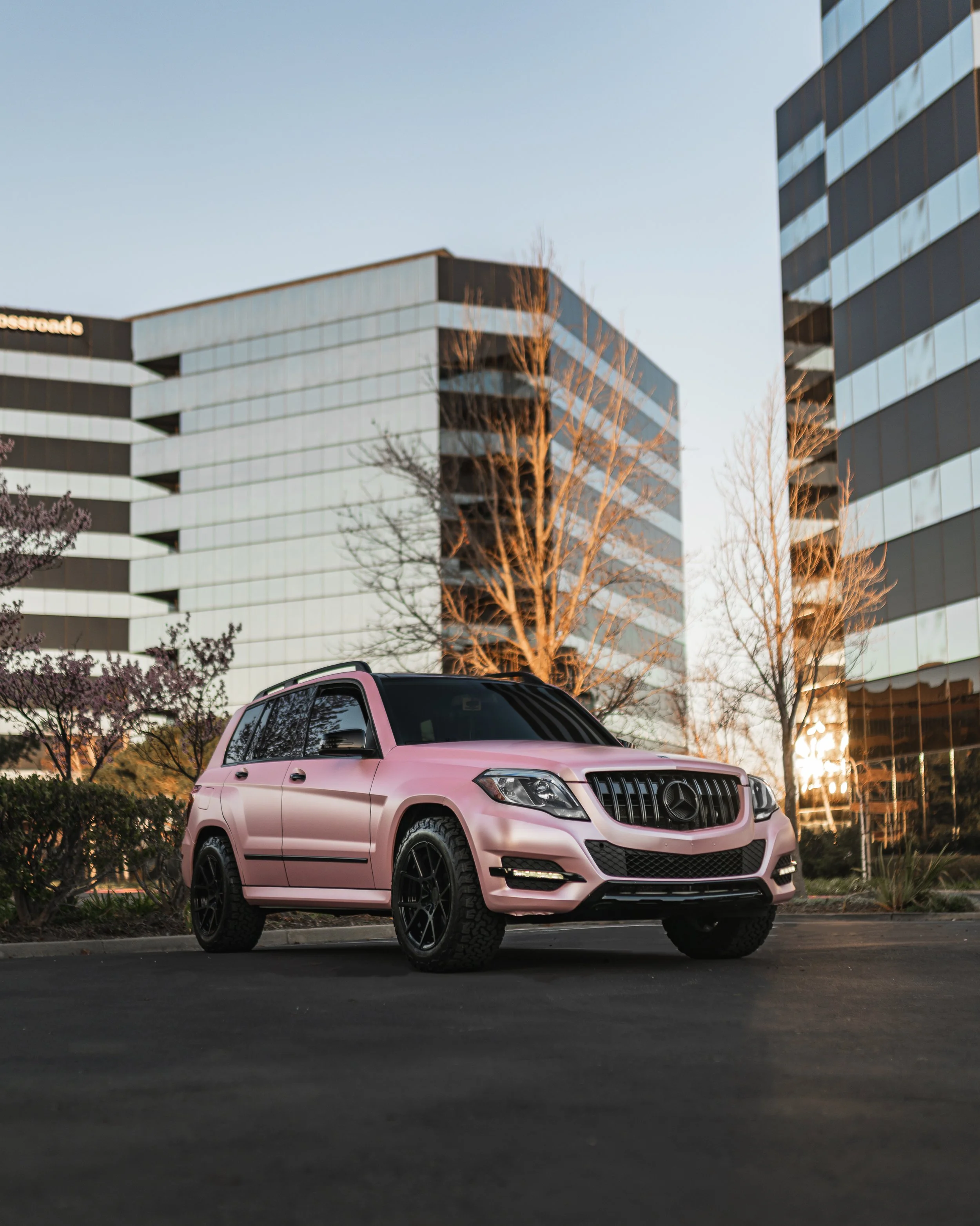 Pink Mercedes-Benz SUV parked on a city street during sunset with office buildings in the background.