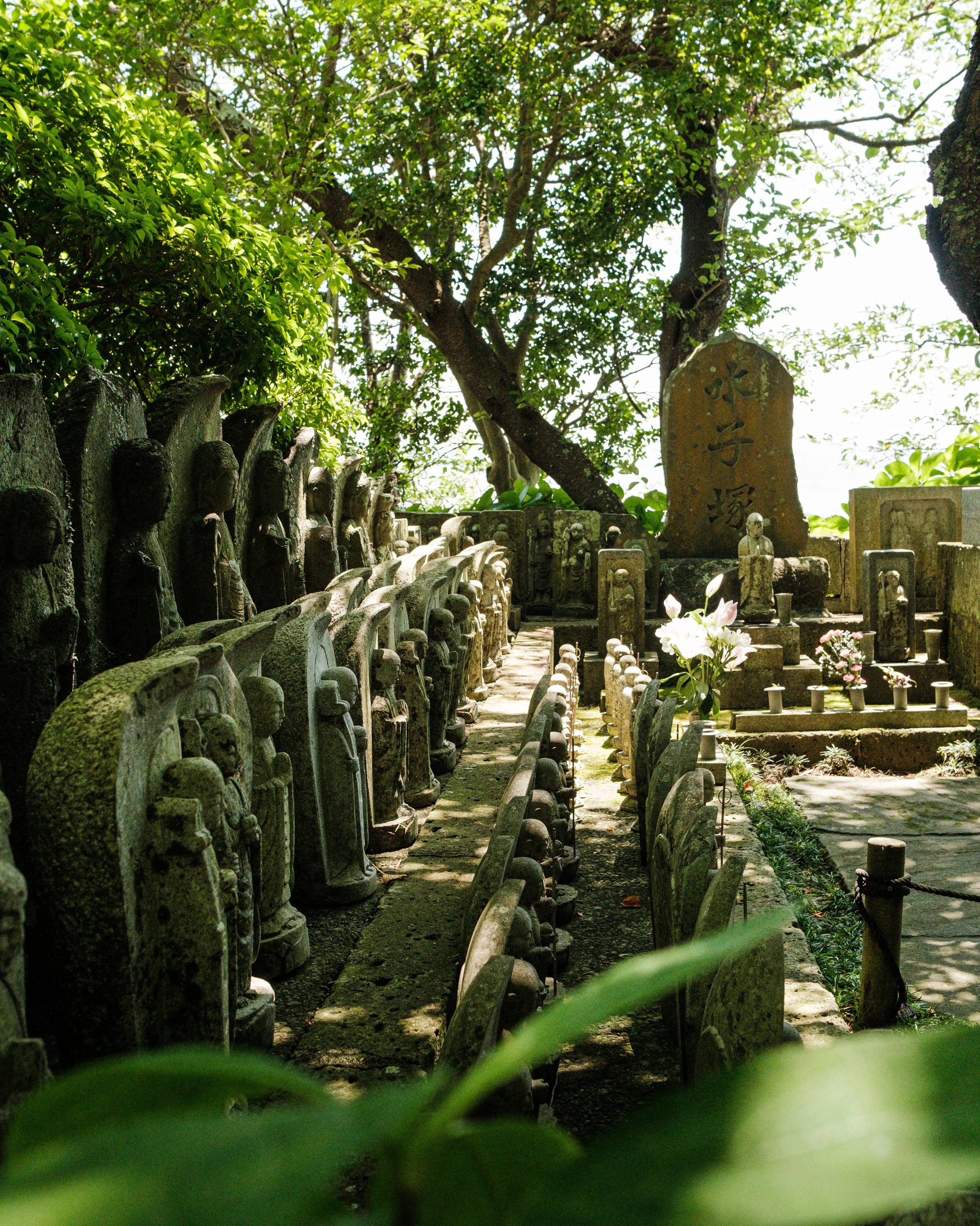 Stone statues and tombstones in a lush, green, outdoor Japanese cemetery or temple yard surrounded by trees and greenery.
