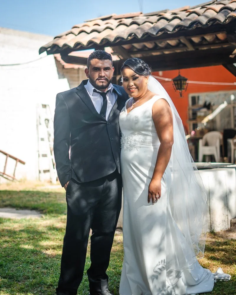 A bride and groom posing outdoors, with the groom in a black suit and the bride in a white wedding gown and veil.