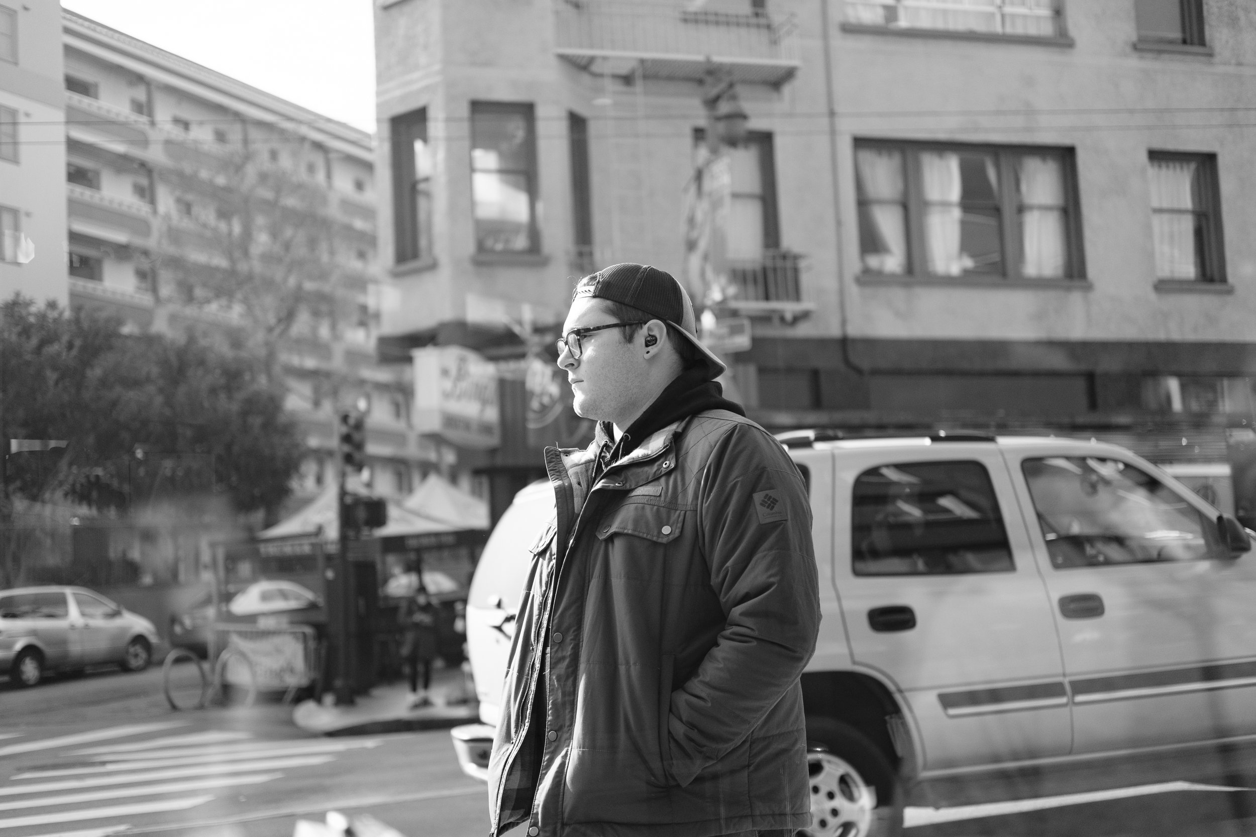 A man with glasses, wearing a backward baseball cap and a jacket, stands on a city street with cars and buildings in the background.