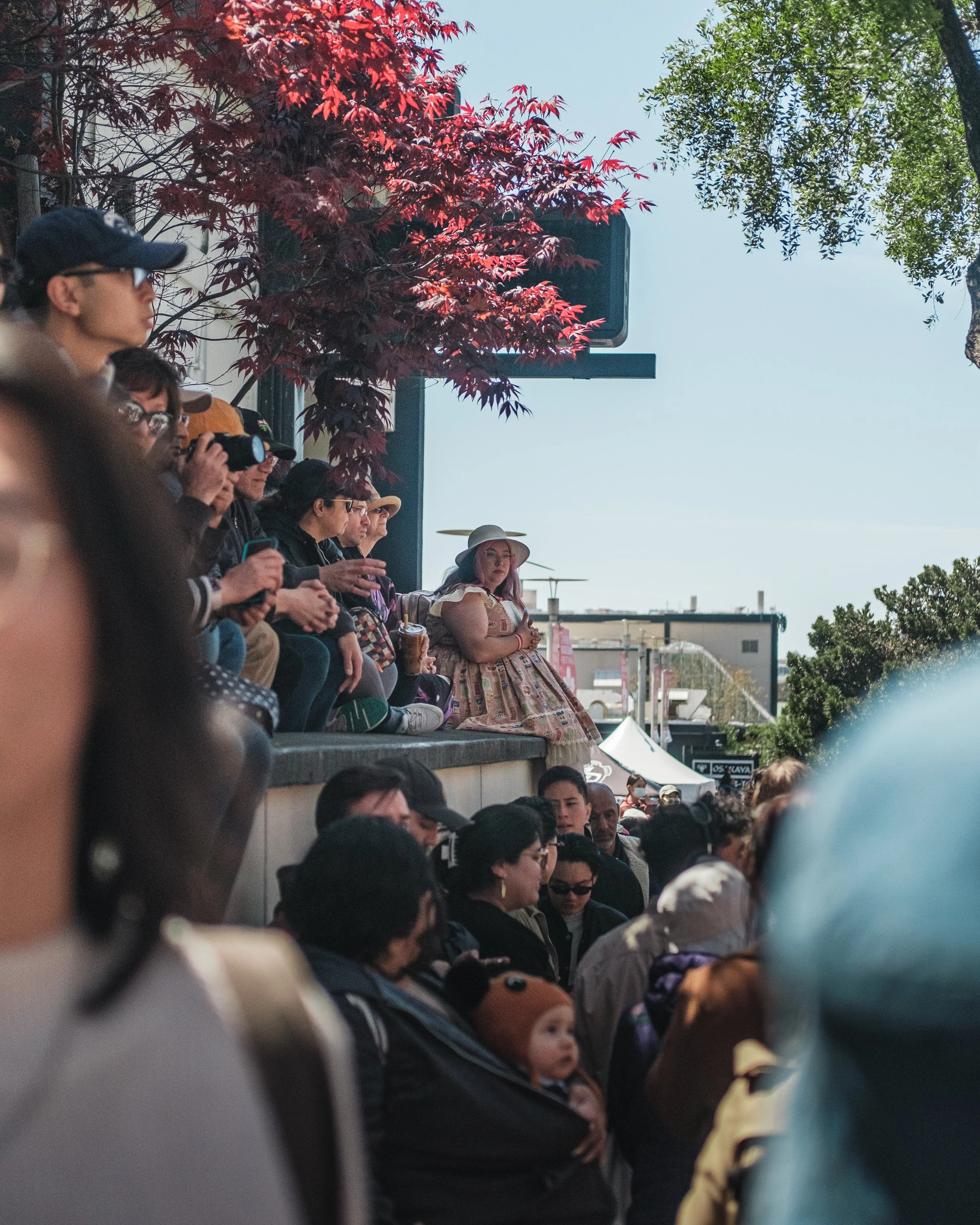 Crowd of people seated and standing outdoors under a tree, with a woman in a floral dress and wide-brimmed hat sitting on a higher platform surrounded by greenery.