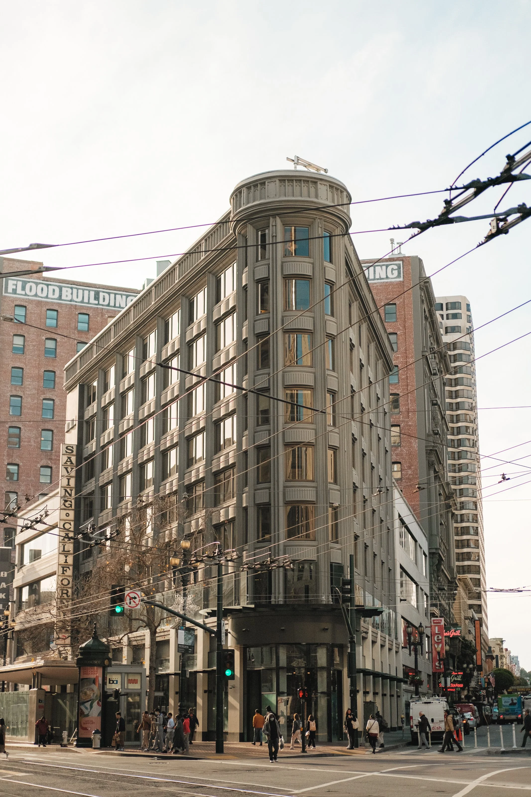 A tall, historic building with rounded corner and decorative details in an urban area with trolley wires overhead and pedestrians on the street.