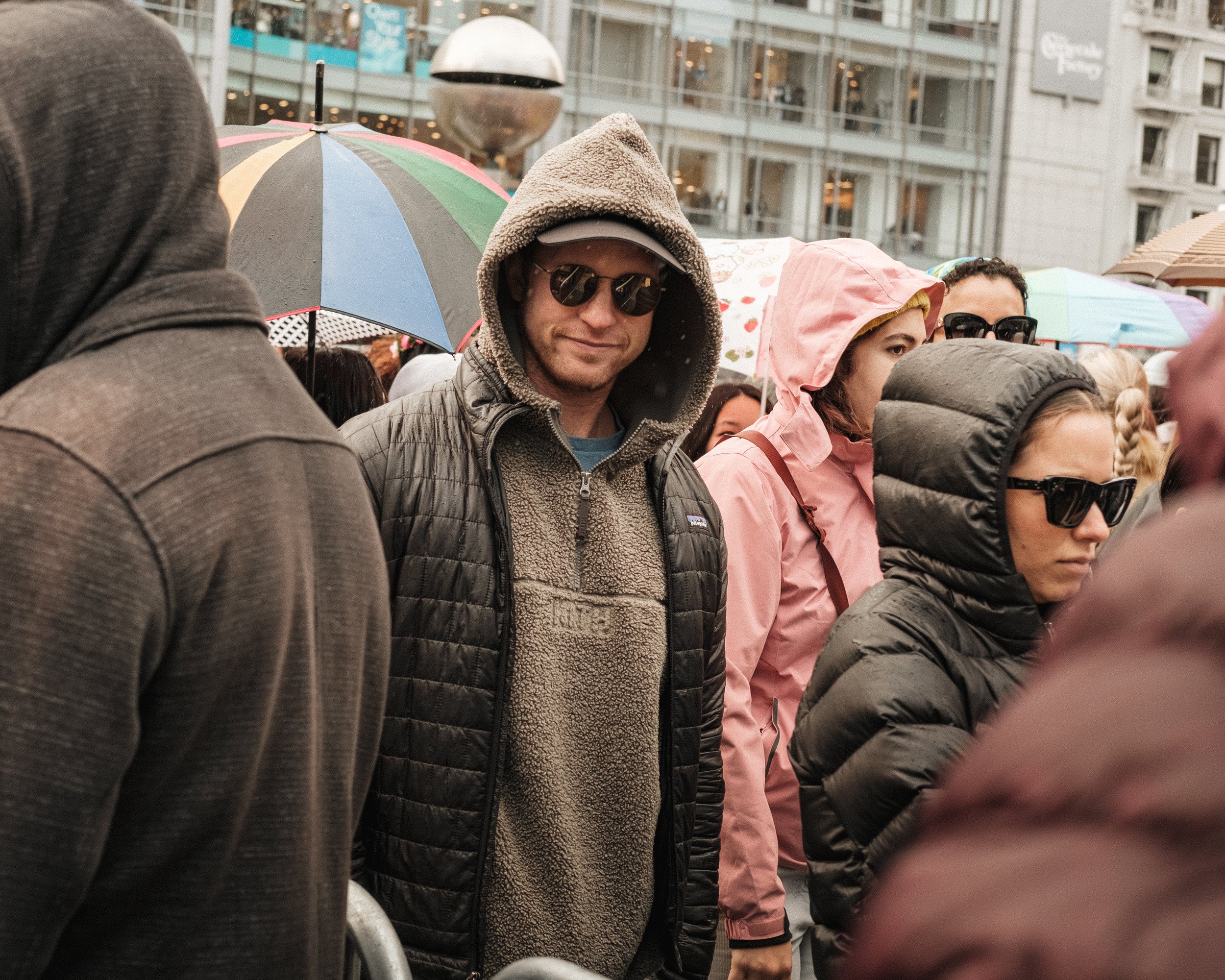 A crowd of people is gathered outdoors on a rainy day, many holding or under colorful umbrellas, with a modern city building in the background. In the center, a man wearing sunglasses, a hoodie, and a fleece jacket is looking at the camera.