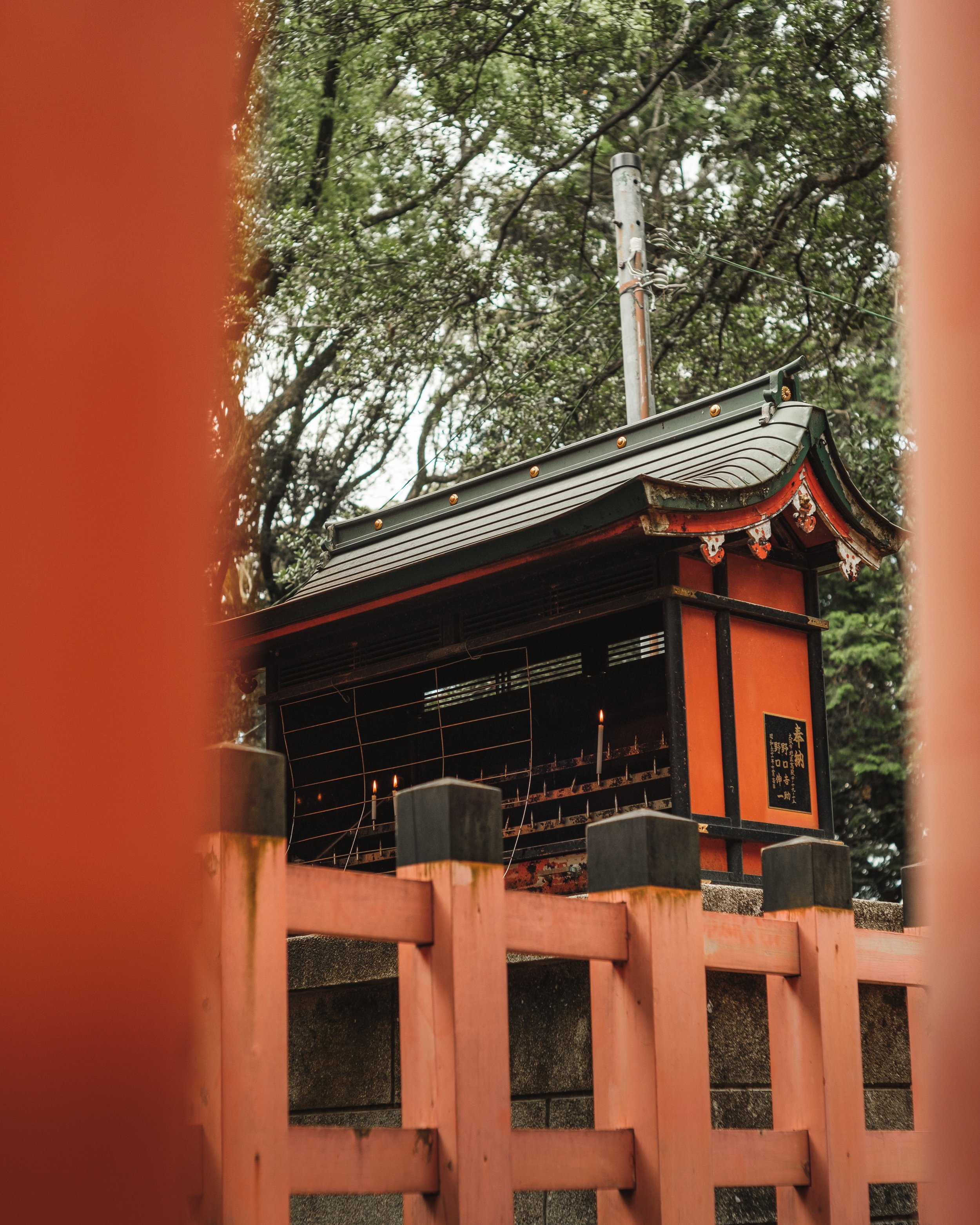 Small traditional Japanese shrine or altar with lit candles, surrounded by a pink fence, with trees and power lines in the background.