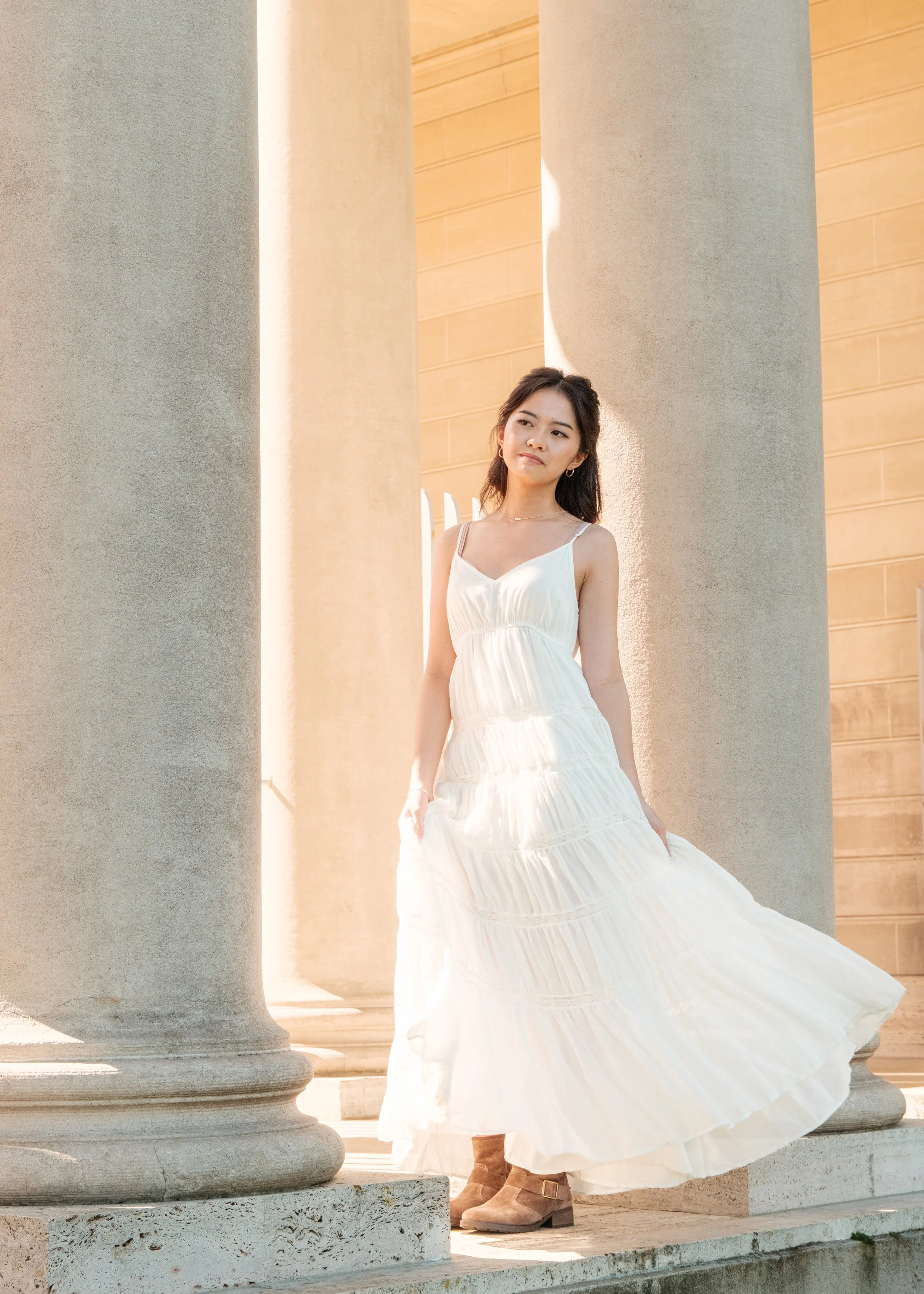 A woman in a white dress standing between large stone columns on a sunlit day.