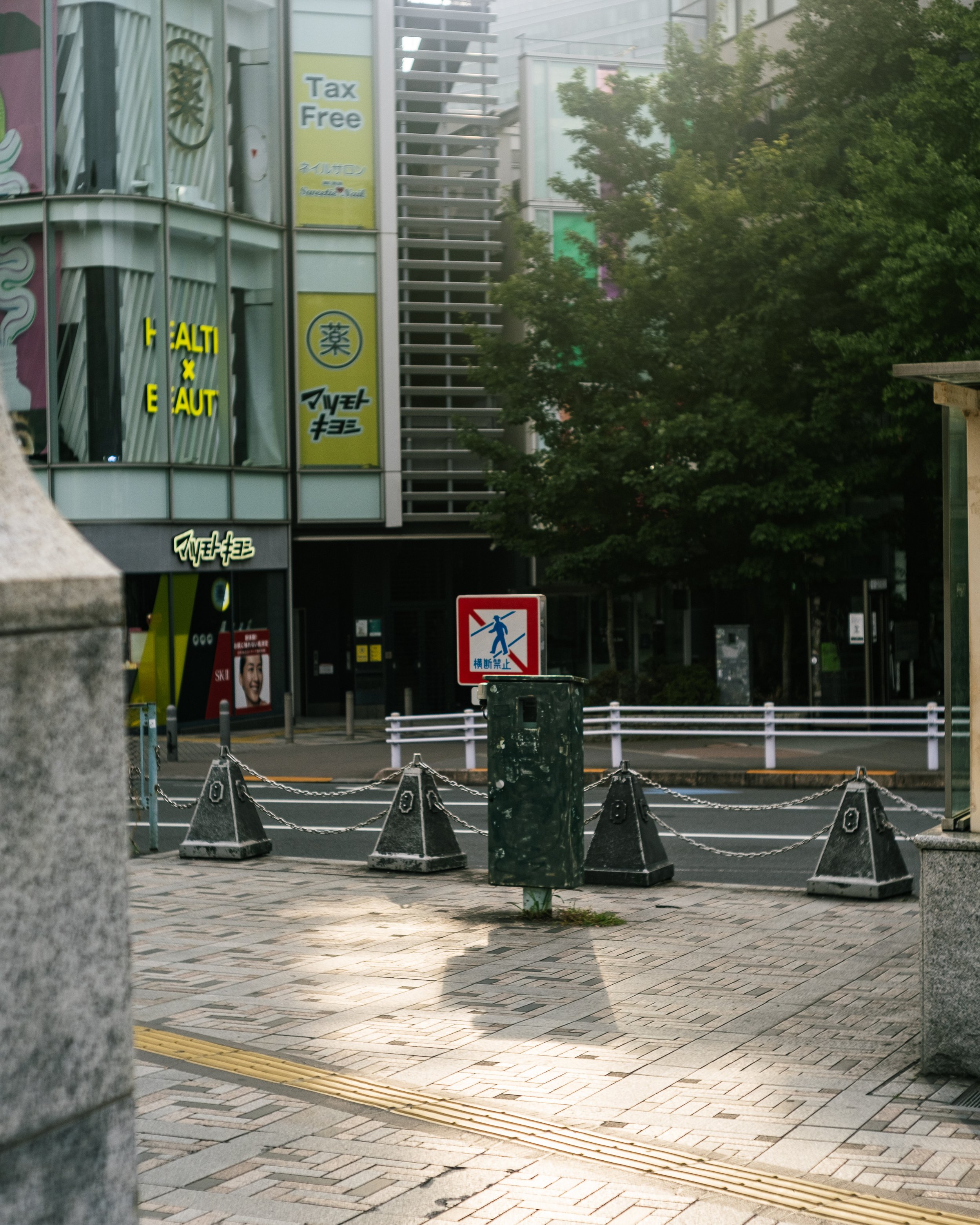 City street scene with a sidewalk, chain barriers, a sign with a red slash indicating no walking, and buildings with signs and trees in the background.