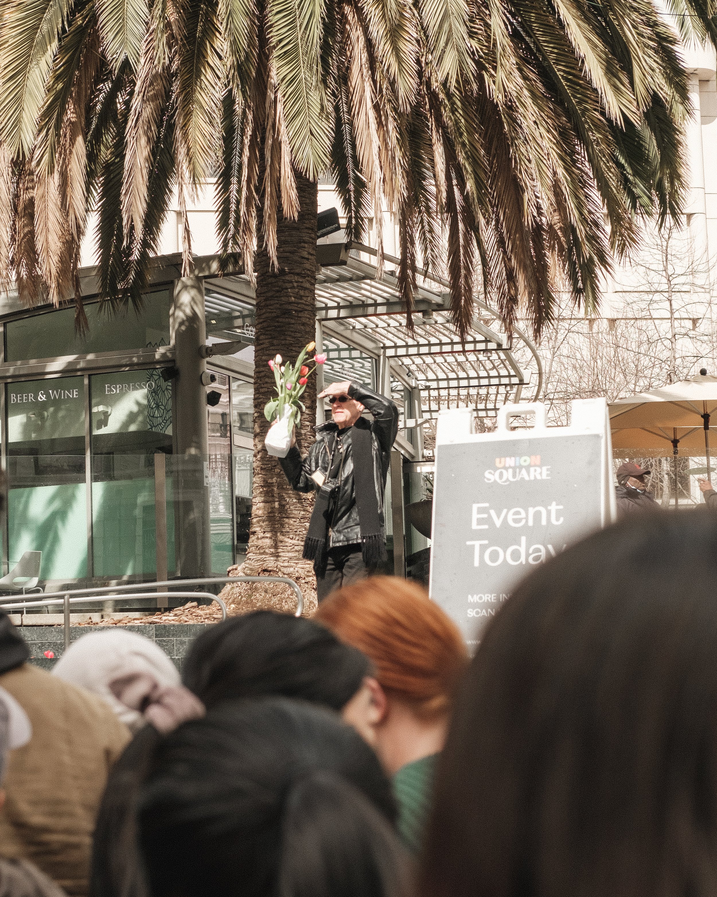 A woman holding a bouquet of tulips on a stage in Union Square during an outdoor event, with a palm tree, a sign reading 'Event Today,' and a crowd of people in the foreground.