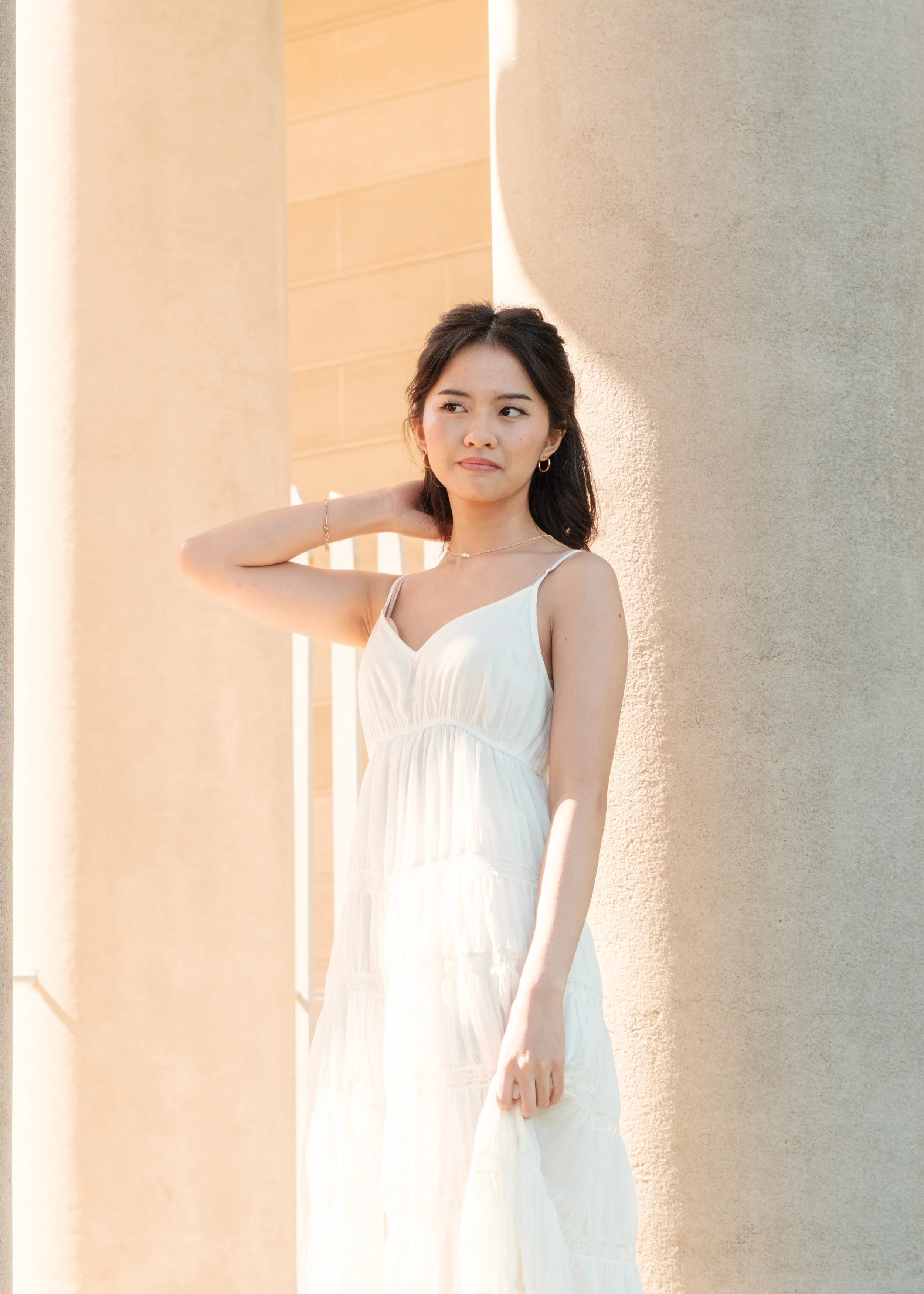 A young woman in a white dress standing outdoors between beige columns during sunset.