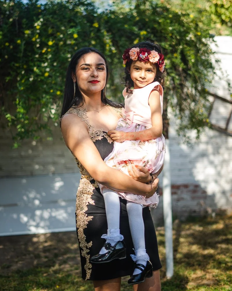 A woman holding a young girl outdoors in front of greenery and a white fence, both smiling and dressed elegantly.