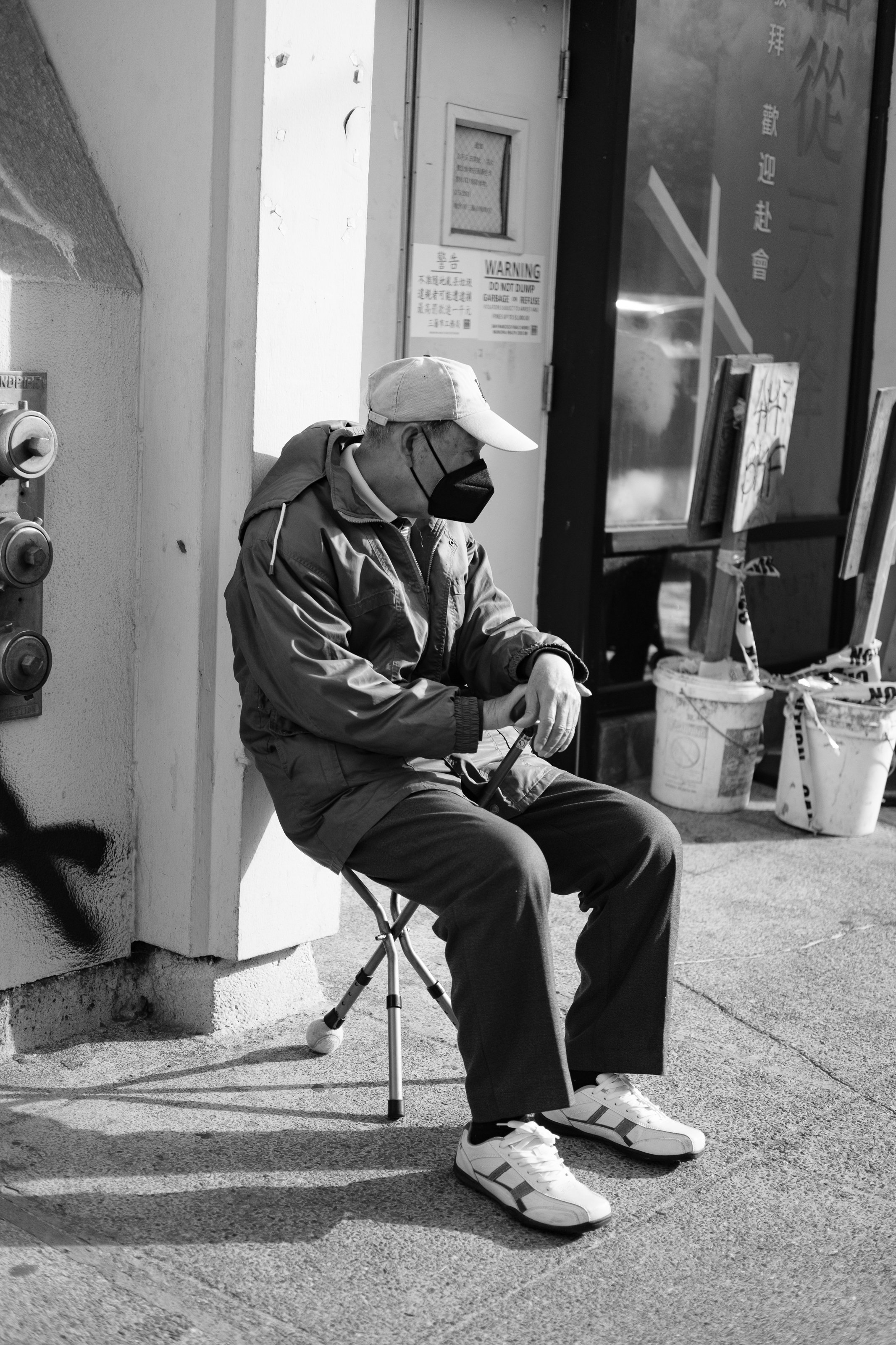 A man wearing a face mask, cap, and casual jacket sits on a folding chair on a sidewalk, looking at his phone with graffiti, warning signs, and buckets in the background.