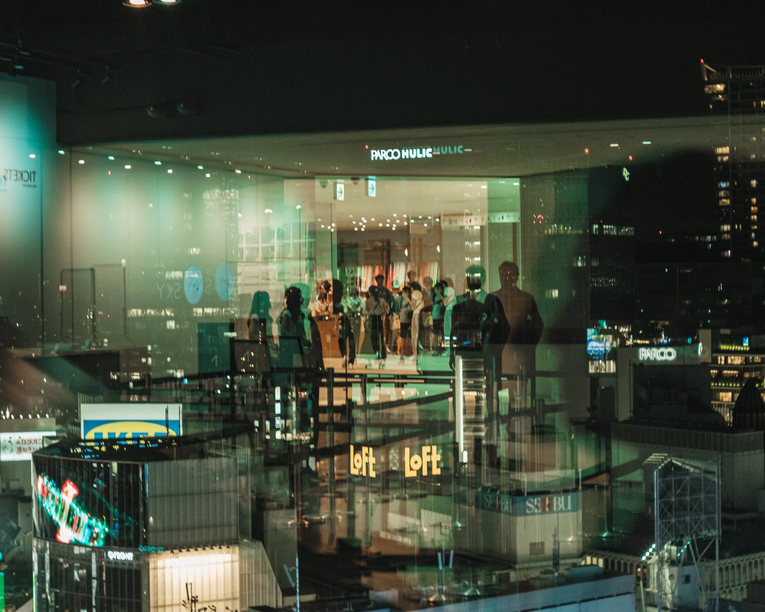 Nighttime view of a rooftop shopping mall with people inside, city buildings with illuminated signs, and reflections on the glass exterior.