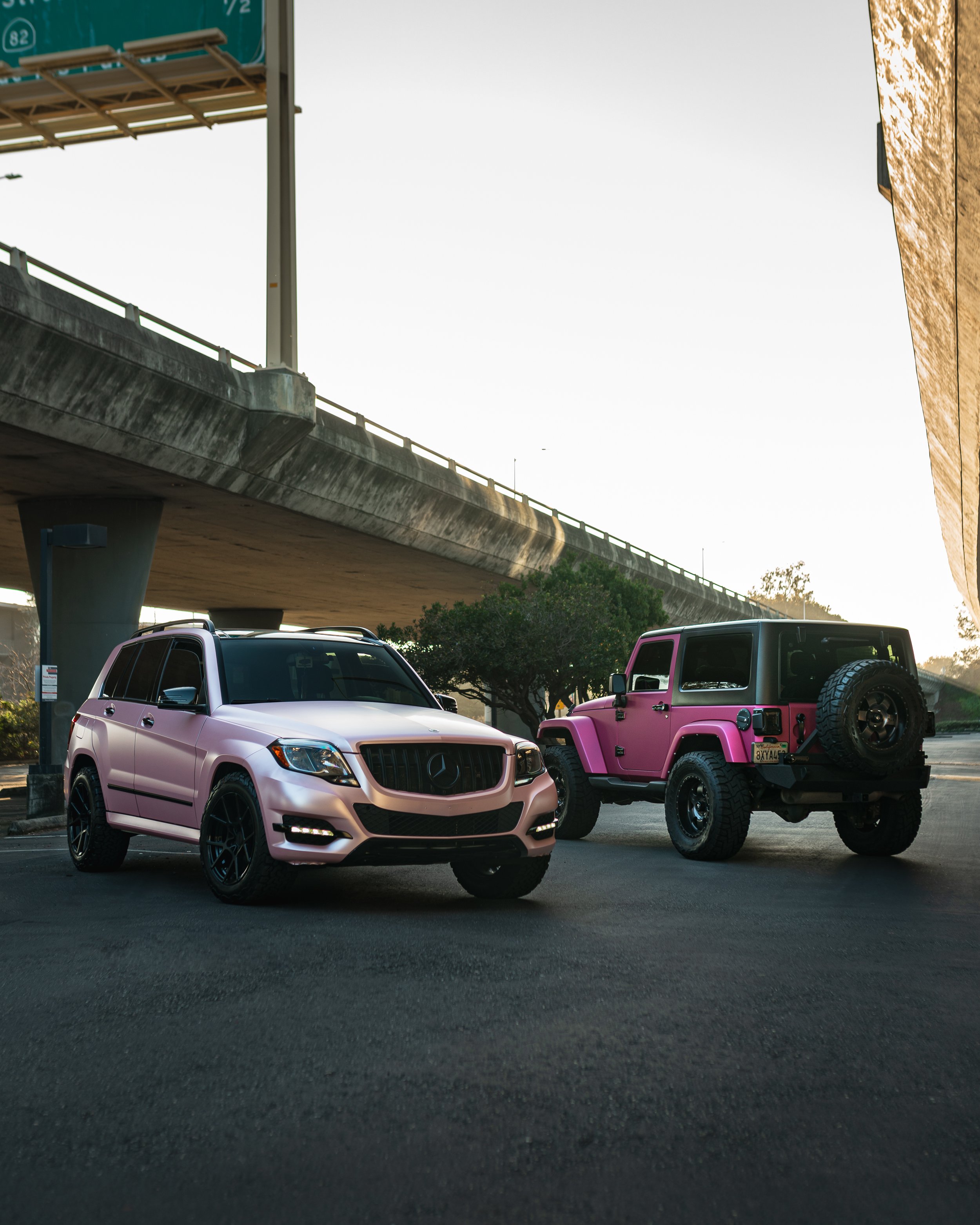 A pink Mercedes-Benz SUV and a pink off-road Jeep parked under an overpass at sunset.