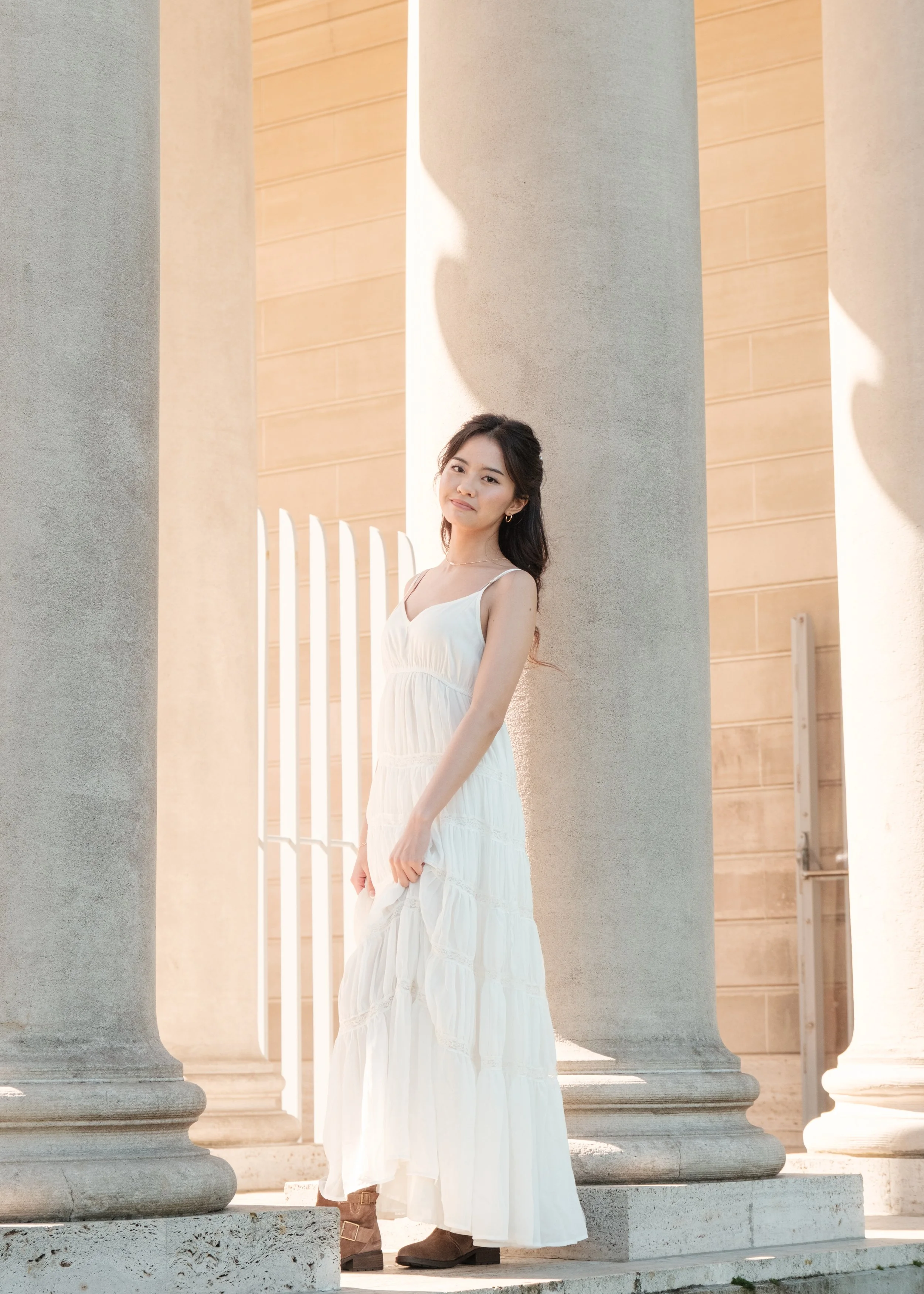 A young woman in a white dress standing between large stone columns outside, with sunlight casting shadows on the columns.