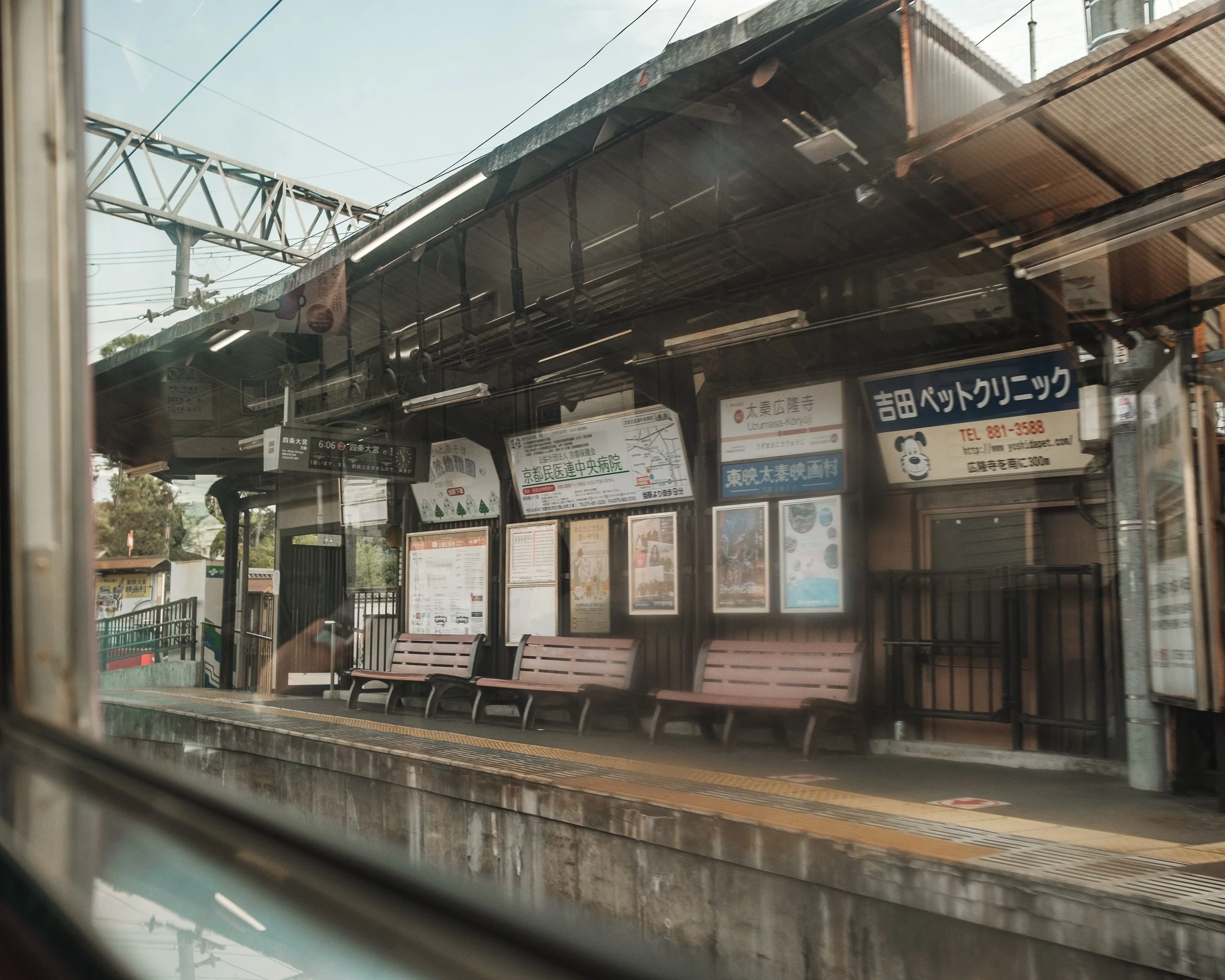 View of an empty train station platform taken from inside a train, with benches, signs, and overhead electrical wires visible.