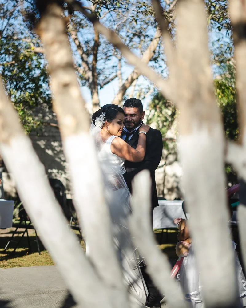 A bride and groom embracing during their wedding, seen through tree branches with greenery in the background.