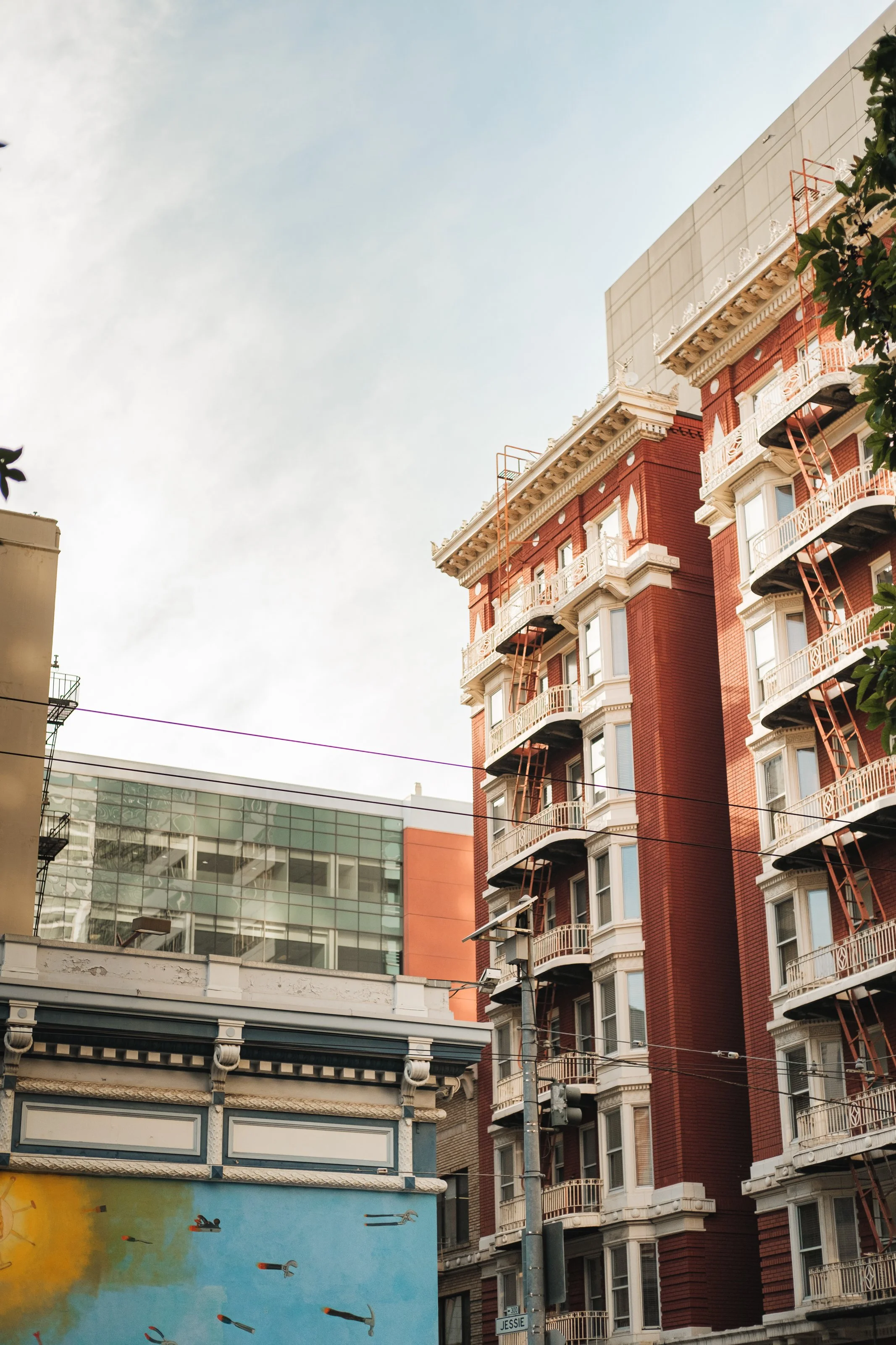 A city street scene featuring a multi-story red brick apartment building with white decorative accents, fire escapes on the balconies, a modern glass building in the background, and a painted wall with flying rocket ship graffiti in the foreground.