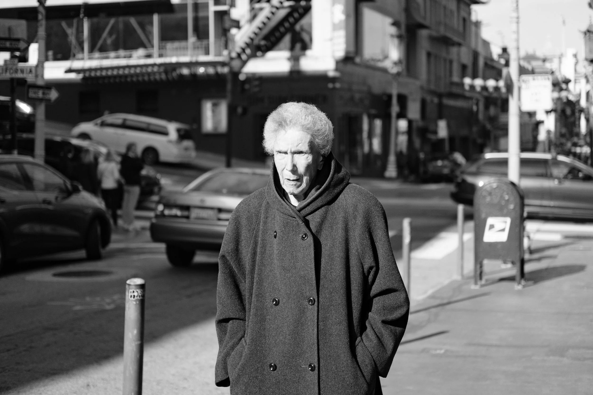 An elderly woman with white hair, wearing a dark coat, walking outdoors on a city street.