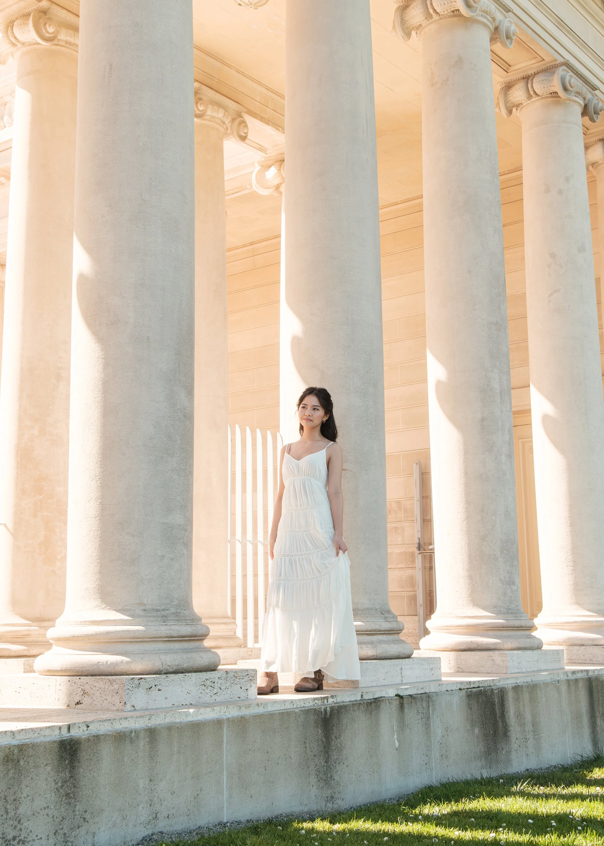 A woman in a white summer dress standing among large white classical columns of a building, with sunlight casting shadows.