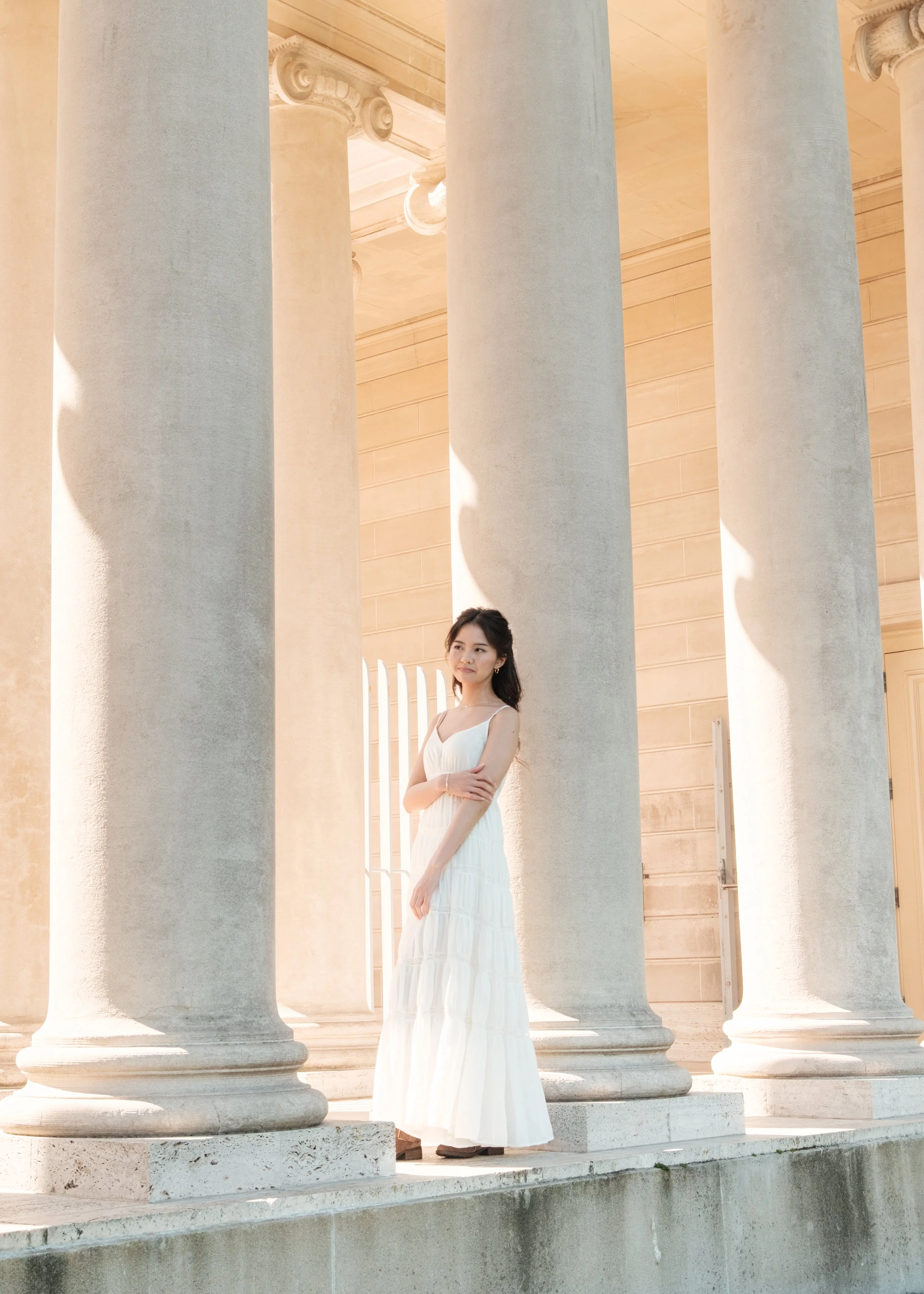 A woman in a white dress standing among large classical columns in sunlight.