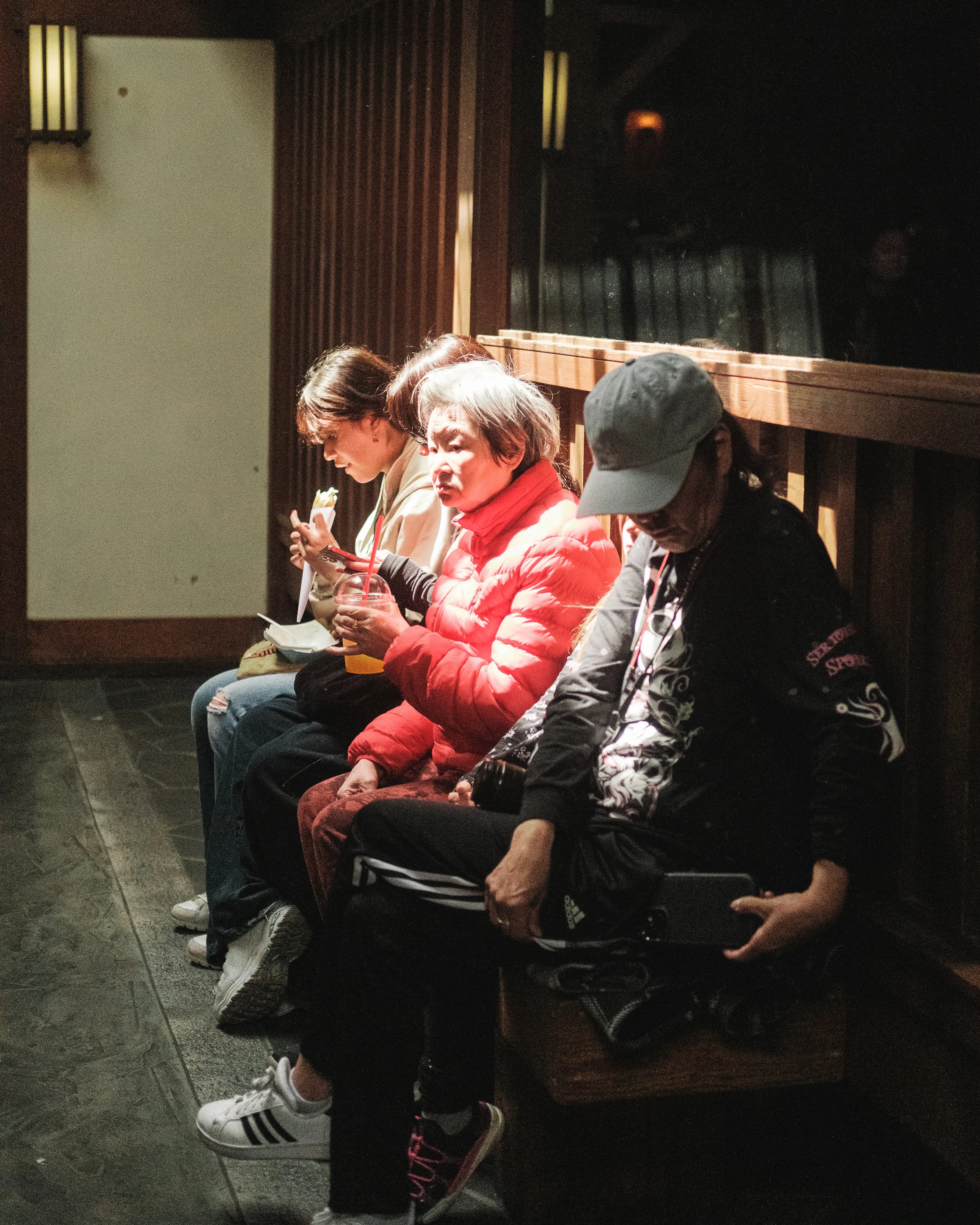 Four women sitting on a wooden bench, one in a red jacket holding a drink, another in a beige jacket looking at her phone, a third in a black cap looking at her phone, inside a dimly lit room with wooden wall paneling.