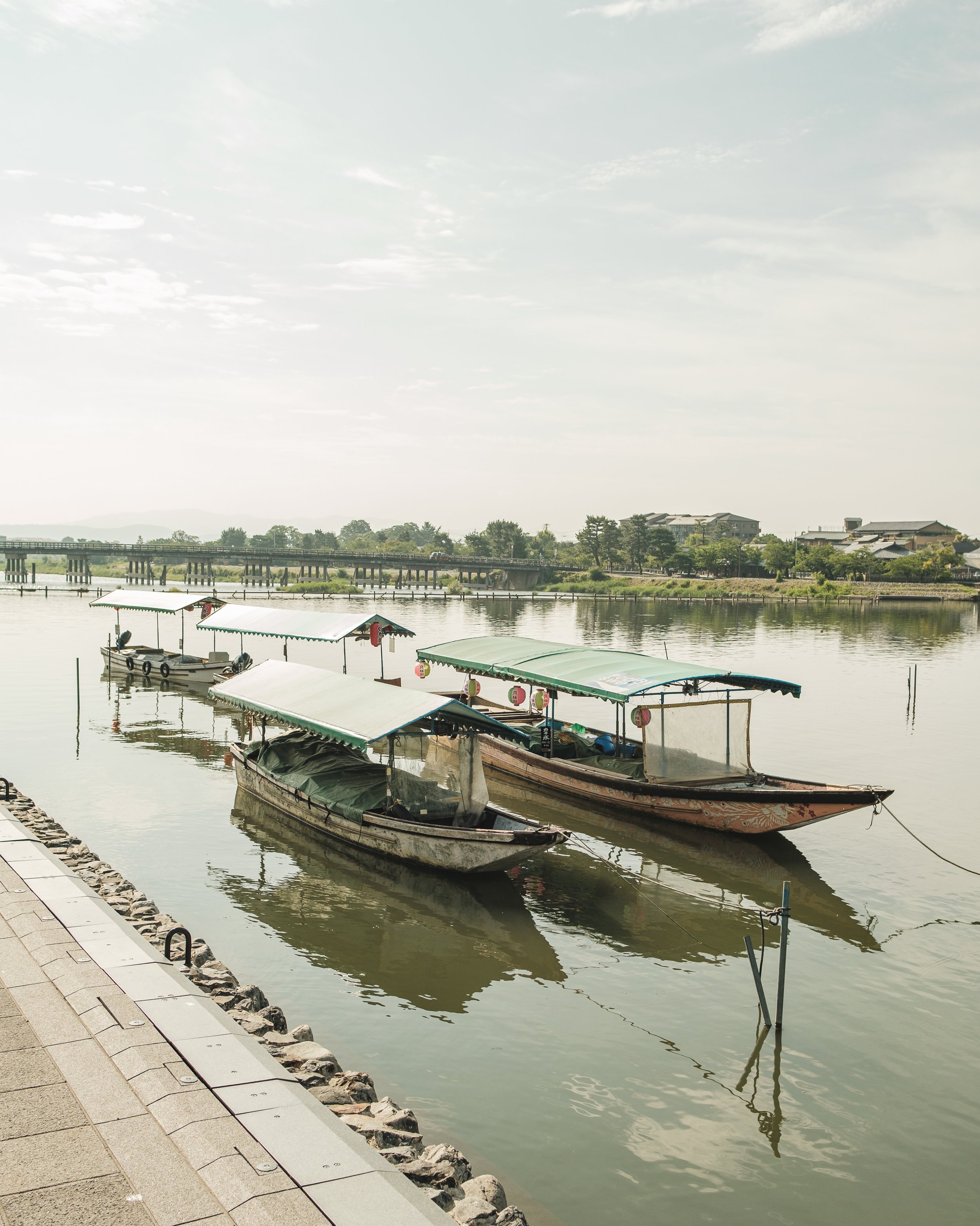 Three small boats docked along a peaceful riverside, with a paved walkway and rocks in the foreground and traditional buildings in the background.