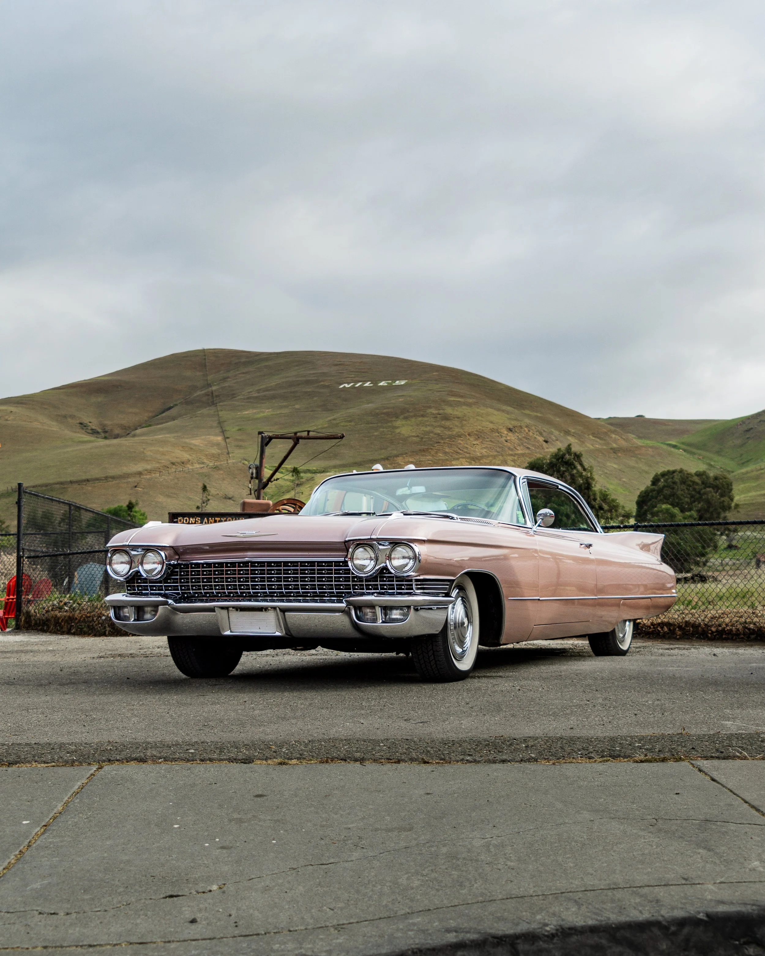 A vintage 1950s Cadillac car parked on the side of a road with grassy hills in the background and a cloudy sky.