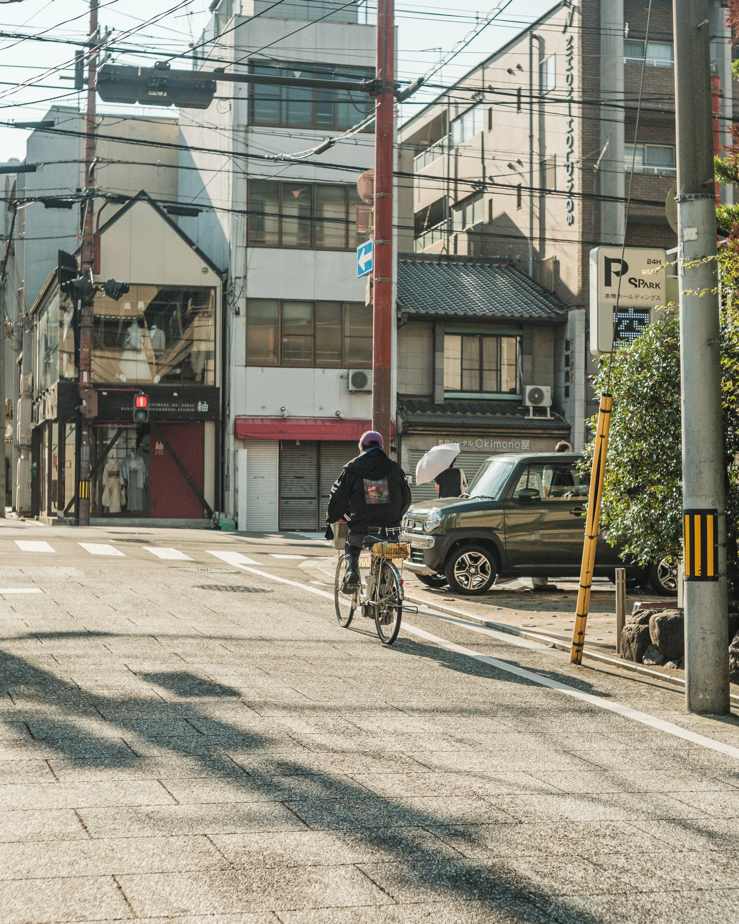 A person riding a bicycle on a city street with buildings, parked cars, and a pedestrian with an umbrella in the background.