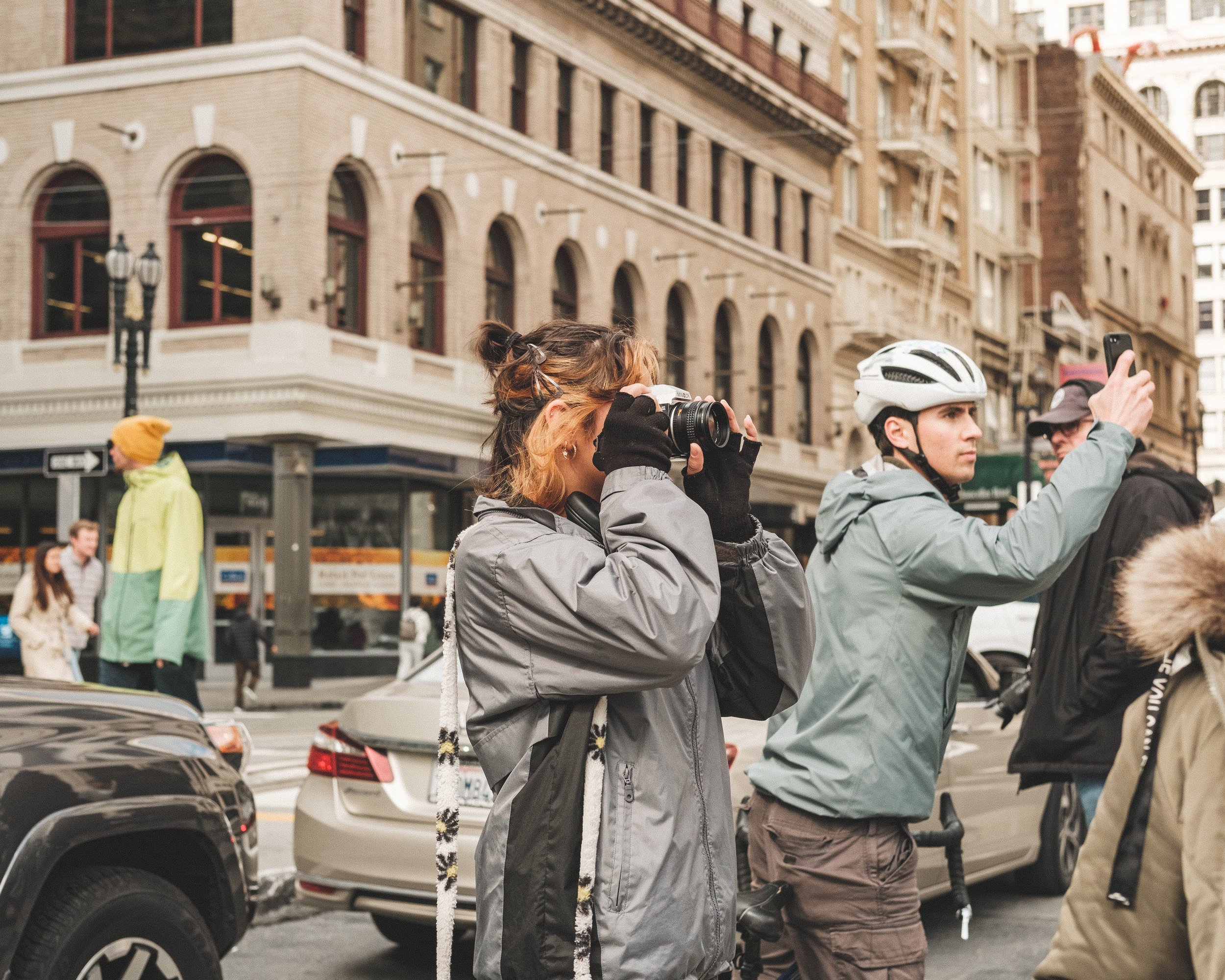Two people, one woman with a camera and one man with a bicycle, standing on a city street. The woman is taking a photo, and the man is using his phone. They are dressed in casual outdoor clothing, with the man wearing a white helmet. The street is bu