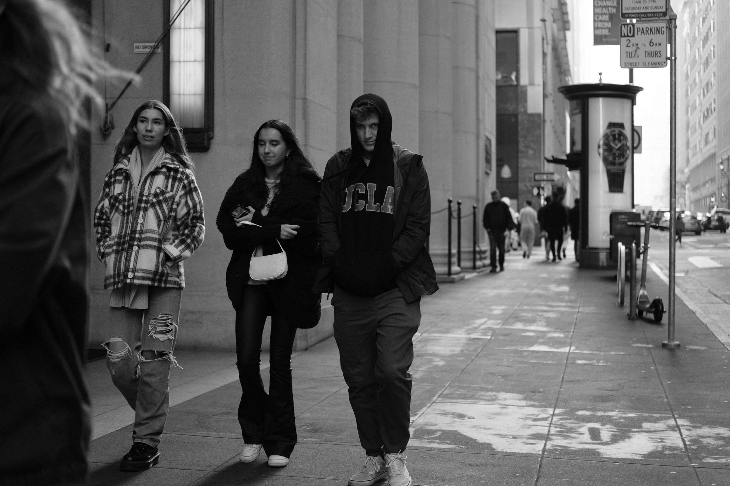 Black and white photo of three young people walking on a city sidewalk. Two women and one man, all dressed casually with the man wearing a UCLA hoodie. The woman on the left is wearing a plaid jacket and ripped jeans, the woman in the middle has long