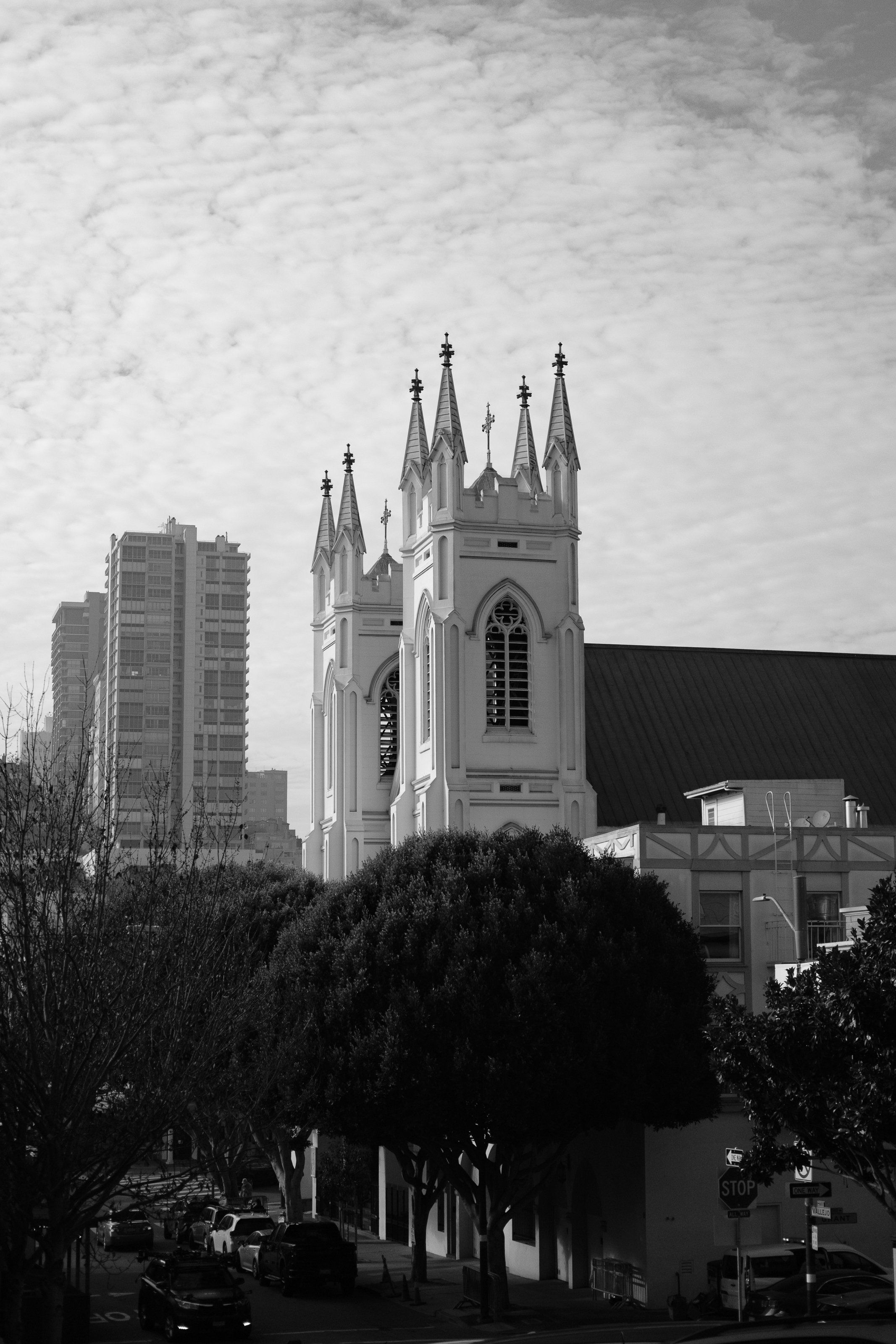 Black and white photo of a church with tall spires, surrounded by trees and buildings, with a cloudy sky overhead.