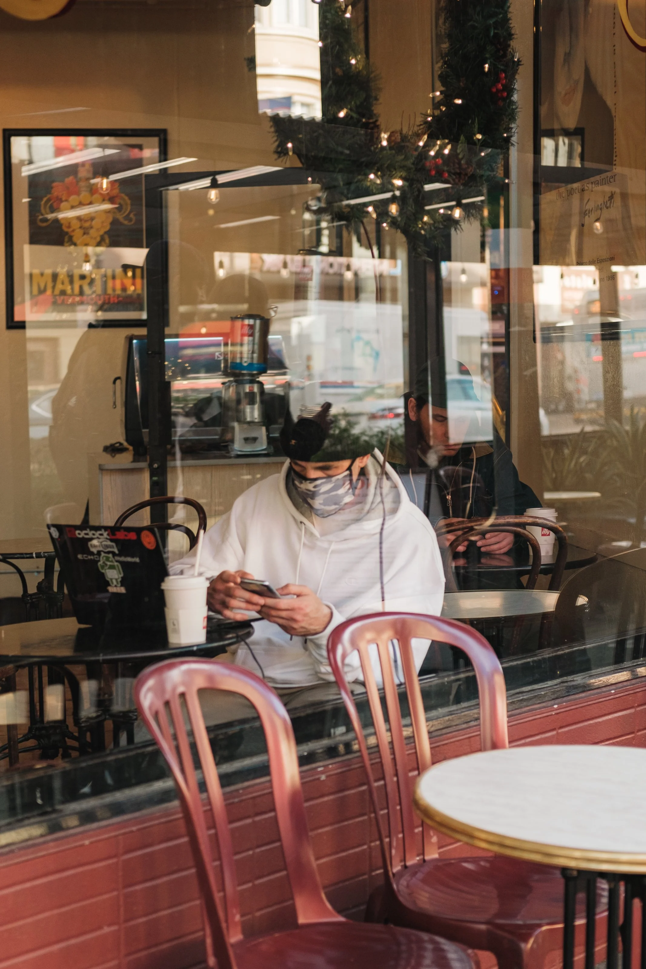 People sitting inside a coffee shop, viewed through a window with reflections. One person is wearing a white hoodie and face mask, using a smartphone. There are empty chairs and tables in the foreground and Christmas decorations inside.