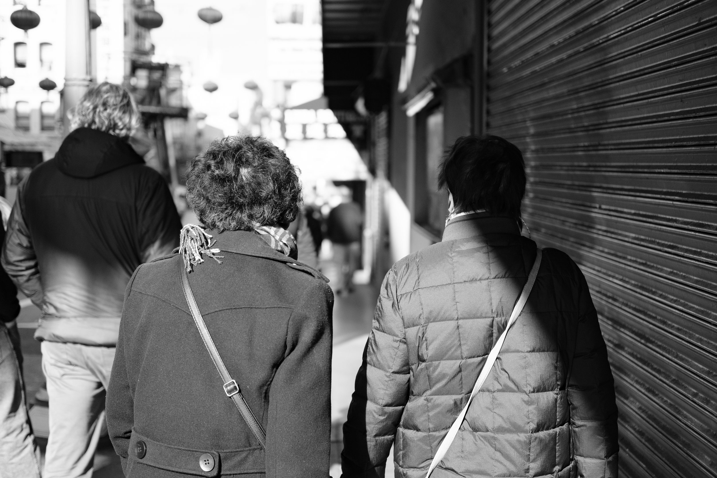 Three people walk down a city sidewalk, with one woman wearing a coat and scarf, and two men in jackets, passing a closed storefront with a shutter.