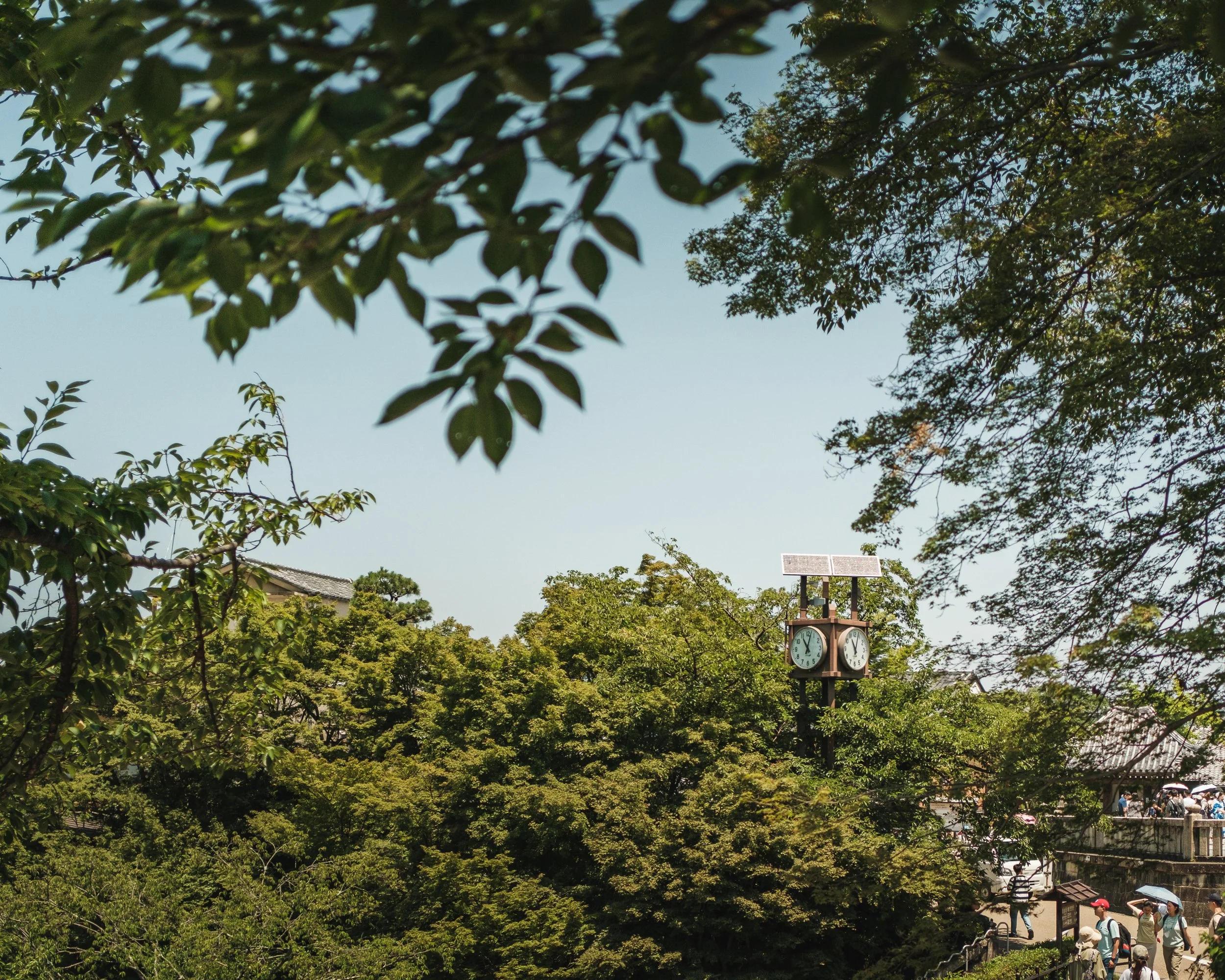 A park scene with lush green trees and a wooden clock tower with two clocks and solar panels on top, located near a crowd of people walking.