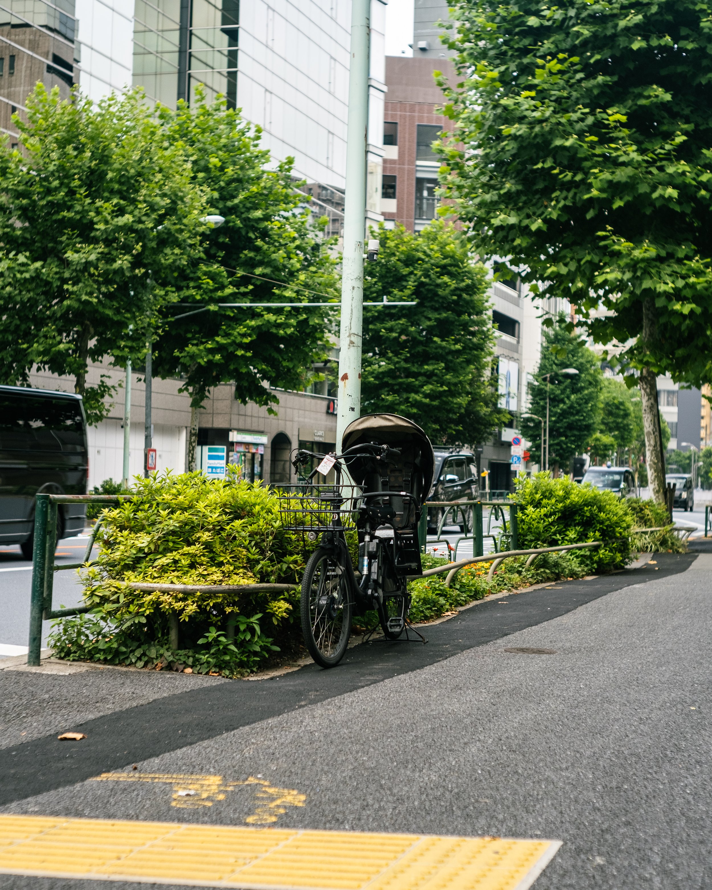 Bicycle with child seat parked on sidewalk near greenery, trees, and low metal railing in an urban area with tall buildings and cars passing by.