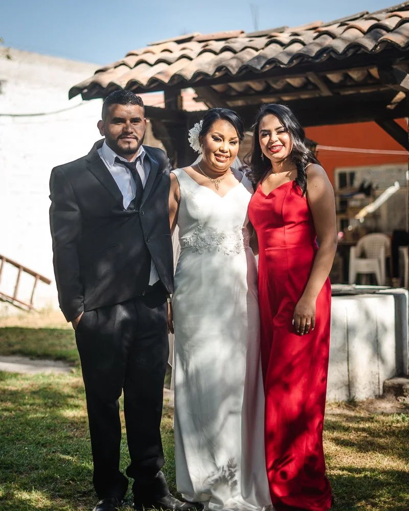 Three people standing outdoors, dressed in formal attire. The man on the left is wearing a black suit, white shirt, and black tie. The woman in the middle is wearing a white wedding dress with floral details. The woman on the right is wearing a red s