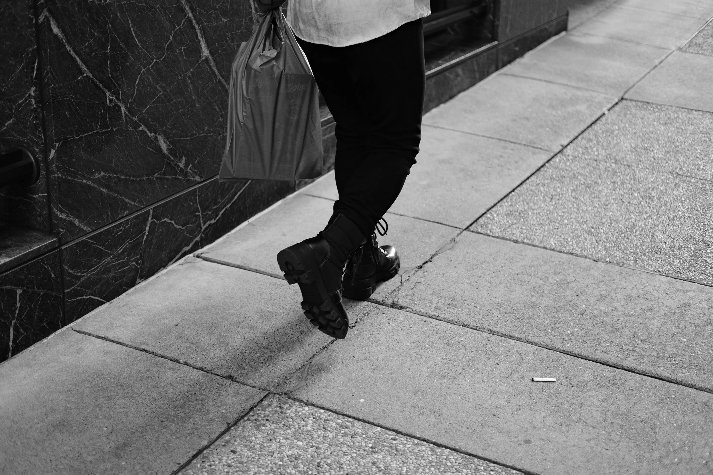 Close-up of a person's legs and feet wearing black boots, black pants, carrying a plastic shopping bag, walking on city sidewalk near building with dark marble exterior.