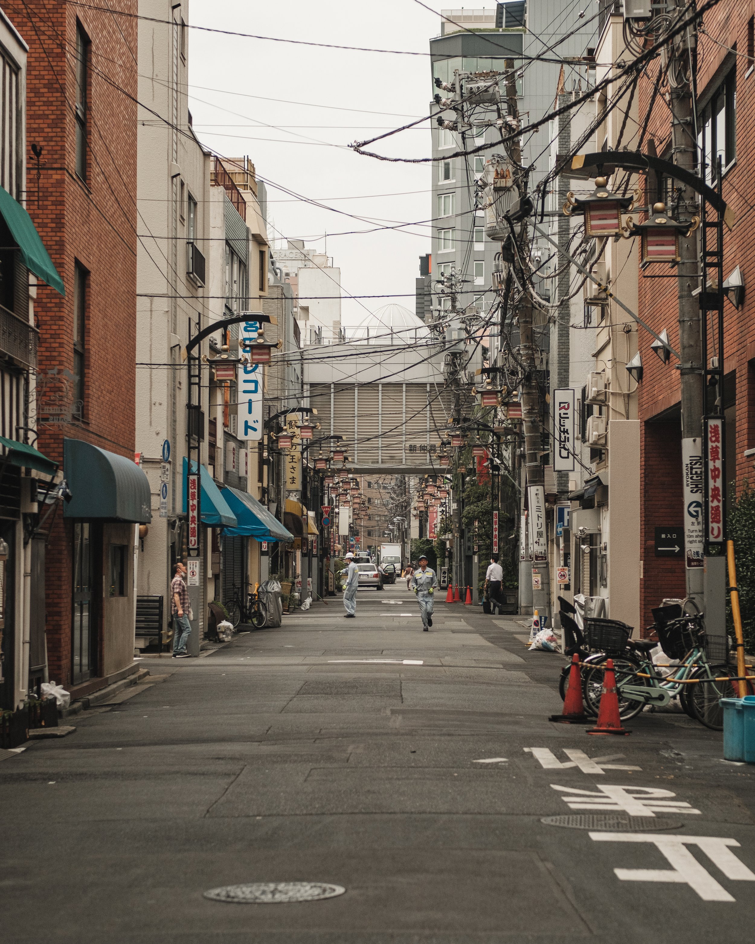 Urban street scene in Japan with buildings, power lines, pedestrians, and bicycles.
